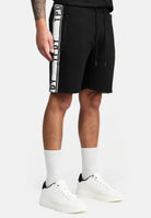 Eine Person trägt trueprodigy Benjamin Logo Shorts mit Reißverschlusstaschen und einem Kordelzug in der Taille, gepaart mit weißen Socken und Turnschuhen. Nur die untere Hälfte des Körpers ist vor einem einfarbigen Hintergrund zu sehen.