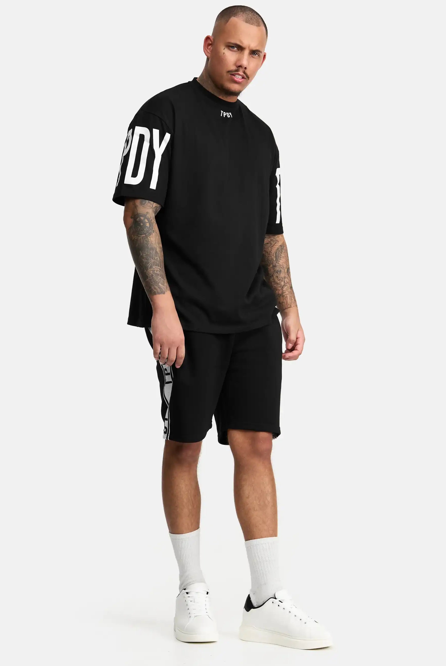 Ein Mann mit kurzen Haaren und Tattoos trägt ein lockeres schwarzes trueprodigy T-Shirt, trueprodigy Benjamin Logo Shorts, weiße Socken und weiße Turnschuhe, während er vor einem schlichten weißen Hintergrund steht.