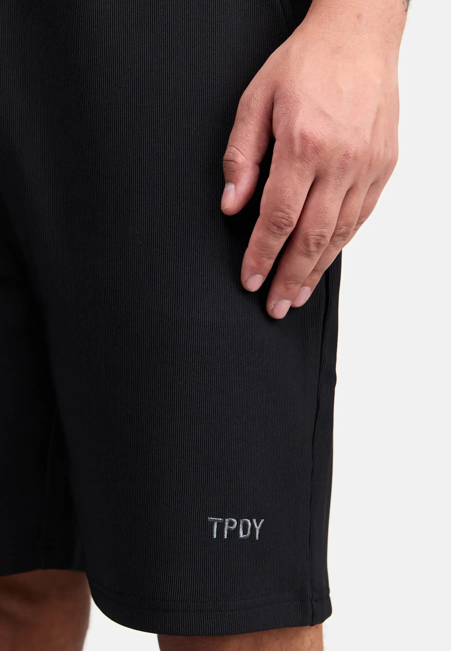 Eine Nahaufnahme zeigt eine Hand, die auf der Samuel Short mit Stickerei von trueprodigy ruht, einer schwarzen Short mit "TPDY"-Stickerei in der Nähe des Saums, vor einem schlichten weißen Hintergrund.