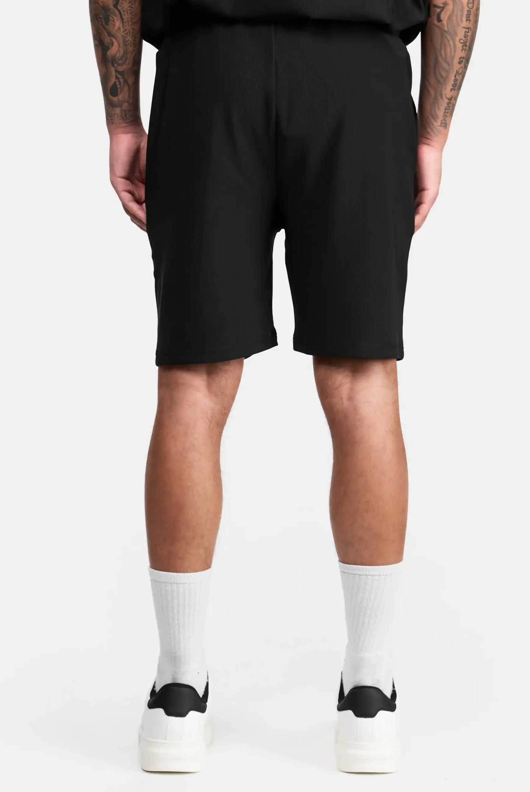 Eine Person steht mit dem Rücken zur Kamera, trägt die schwarze trueprodigy Samuel Short mit Stickerei, ein schwarzes Shirt, weiße Crew-Socken und schwarze trueprodigy Sneaker. Ihre tätowierten Arme hängen entspannt herab vor weißem Hintergrund.