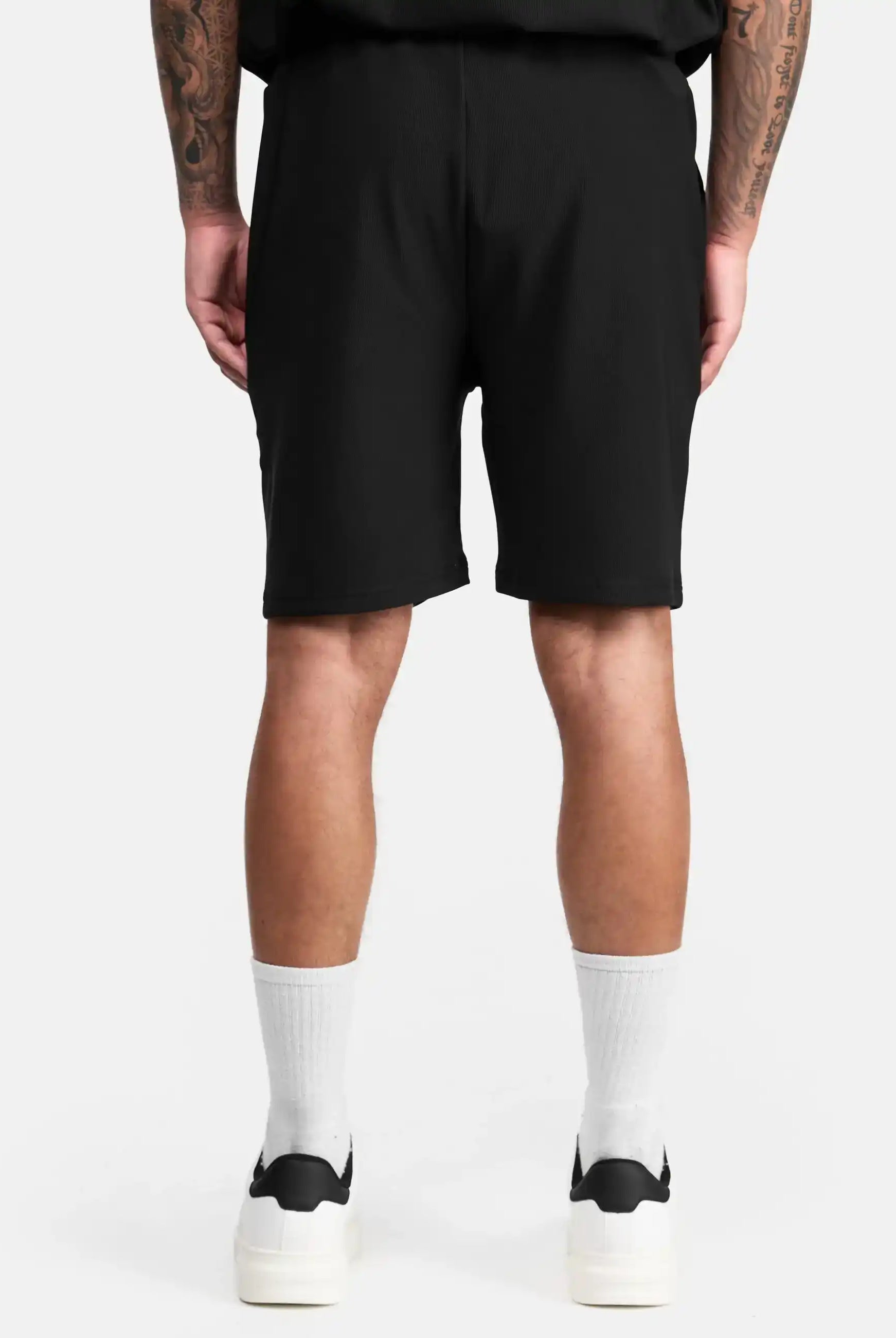 Eine Person steht mit dem Rücken zur Kamera, trägt die schwarze trueprodigy Samuel Short mit Stickerei, ein schwarzes Shirt, weiße Crew-Socken und schwarze trueprodigy Sneaker. Ihre tätowierten Arme hängen entspannt herab vor weißem Hintergrund.