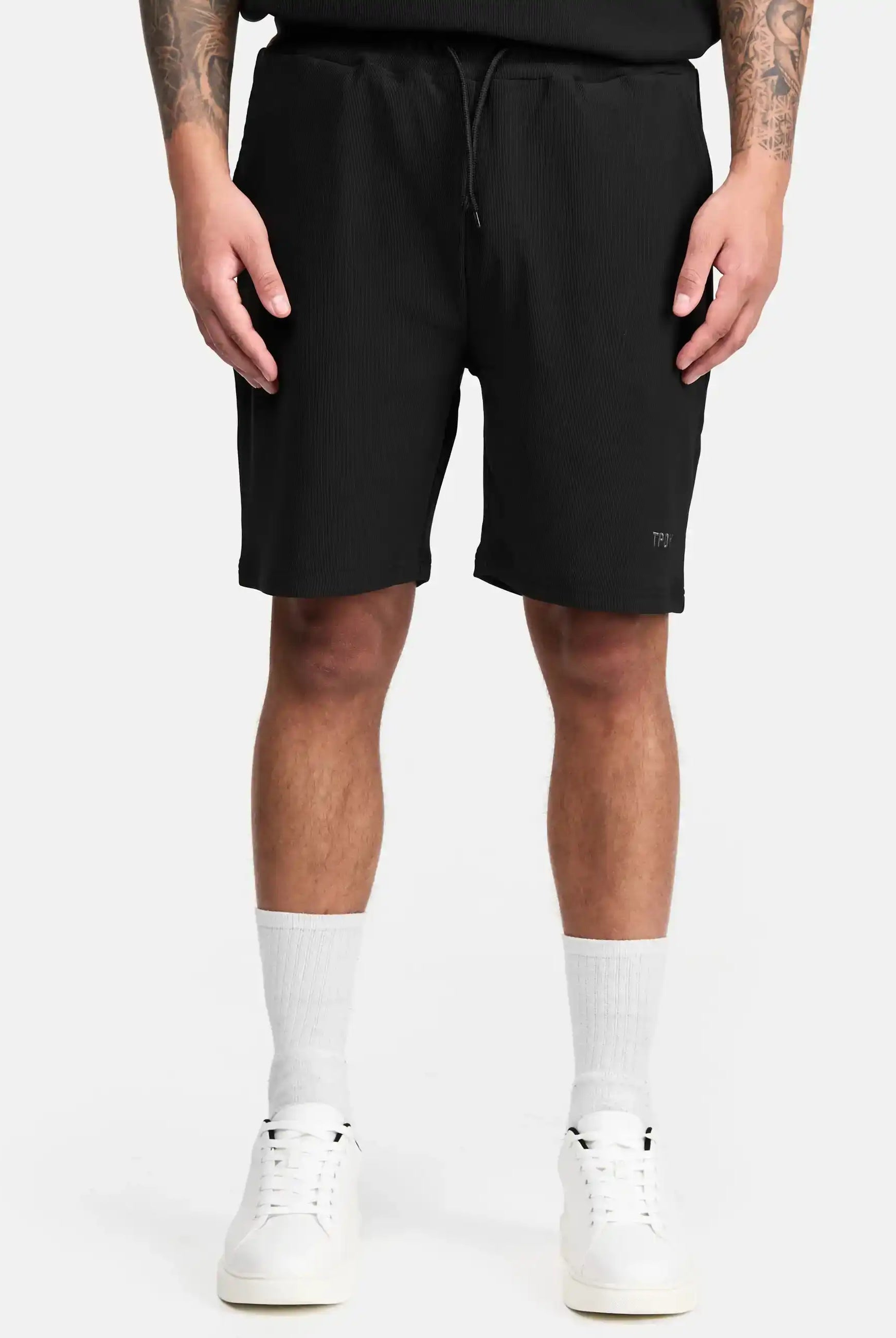Eine Person trägt die Samuel Short mit Stickerei von trueprodigy in Schwarz und ein schwarzes Shirt, dazu weiße Crew-Socken und Sneakers. Die Arme hängen locker an den Seiten, das Foto zeigt sie von den Schultern abwärts.