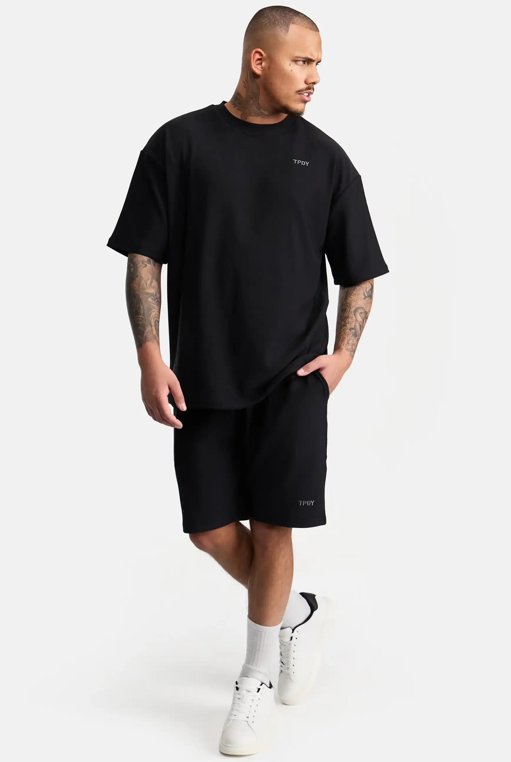 Ein Mann mit kurzen Haaren und Tattoos trägt ein lockeres schwarzes trueprodigy-T-Shirt, Samuel Short mit Stickerei von trueprodigy, weiße Socken und weiße Turnschuhe und steht vor einem schlichten weißen Hintergrund.