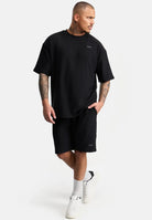 Ein Mann mit kurzen Haaren und Tattoos trägt ein lockeres schwarzes trueprodigy-T-Shirt, Samuel Short mit Stickerei von trueprodigy, weiße Socken und weiße Turnschuhe und steht vor einem schlichten weißen Hintergrund.