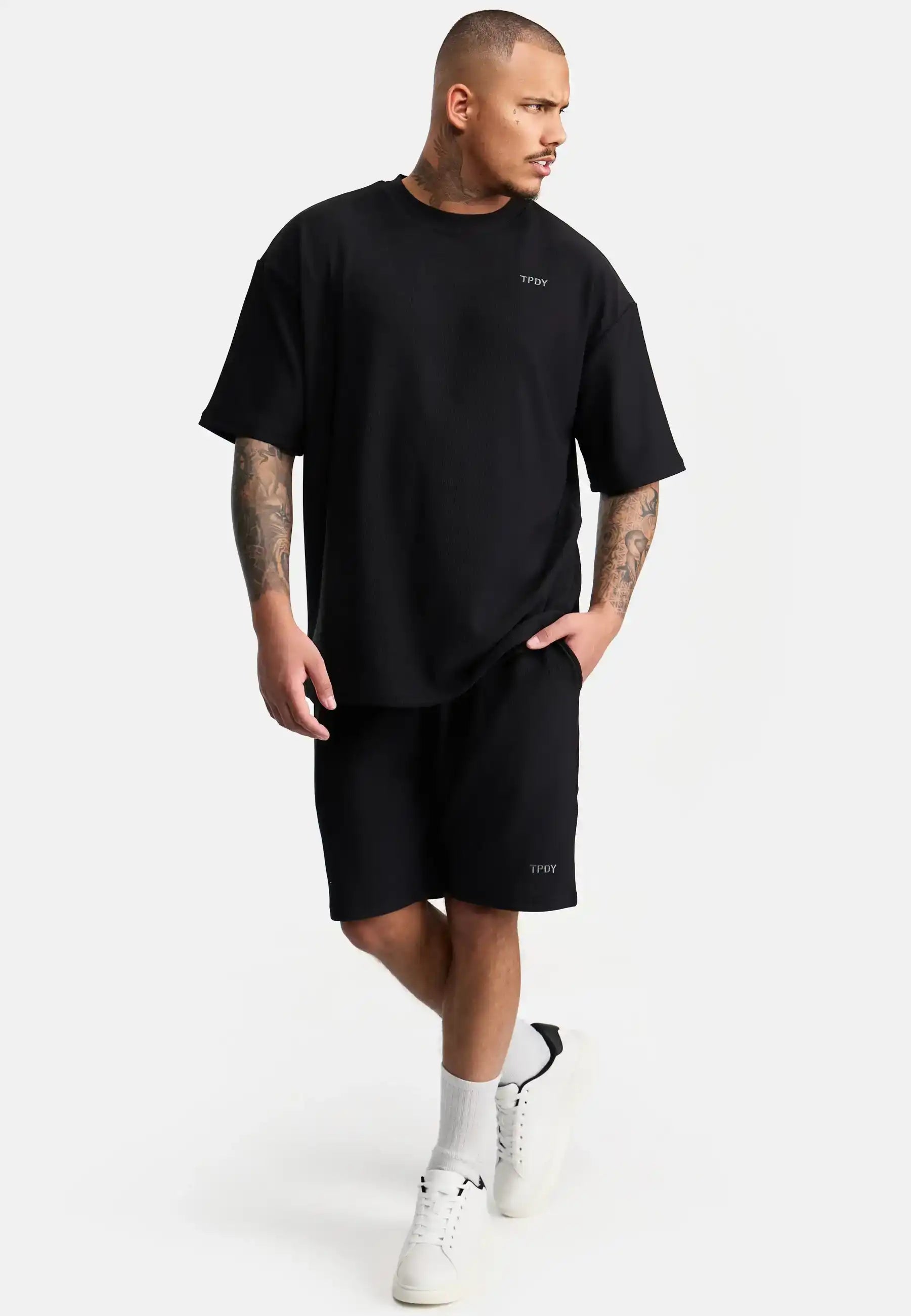 Ein Mann mit kurzen Haaren und Tattoos trägt ein lockeres schwarzes trueprodigy-T-Shirt, Samuel Short mit Stickerei von trueprodigy, weiße Socken und weiße Turnschuhe und steht vor einem schlichten weißen Hintergrund.