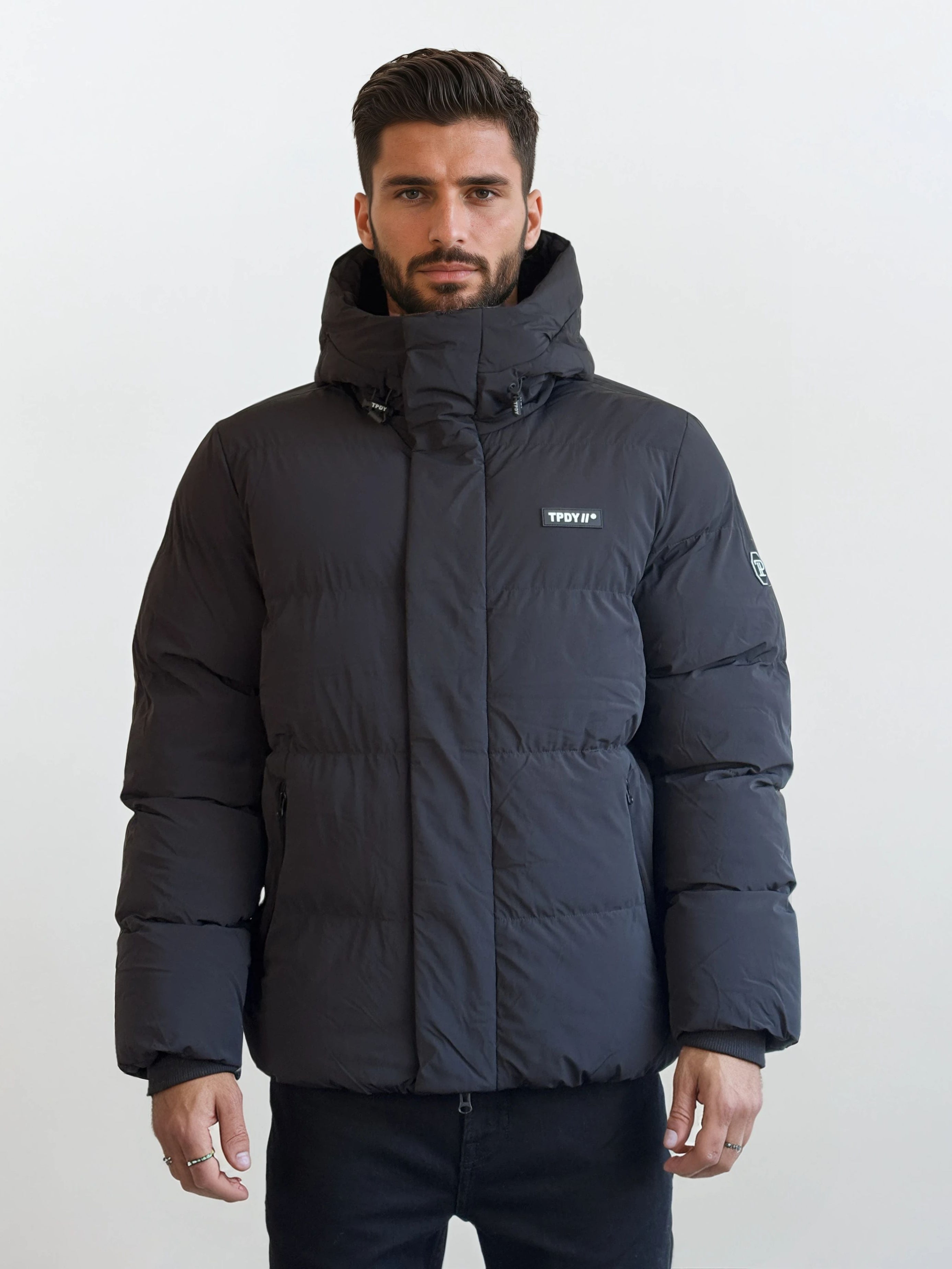 Ein Mann mit kurzen dunklen Haaren und einem Bart trägt eine schwarze Pufferjacke mit Kapuze vor einem schlichten weißen Hintergrund. Die Jacke hat den Logo-Text "Trophy" auf der linken Brust und einen Aufnäher auf dem linken Ärmel.