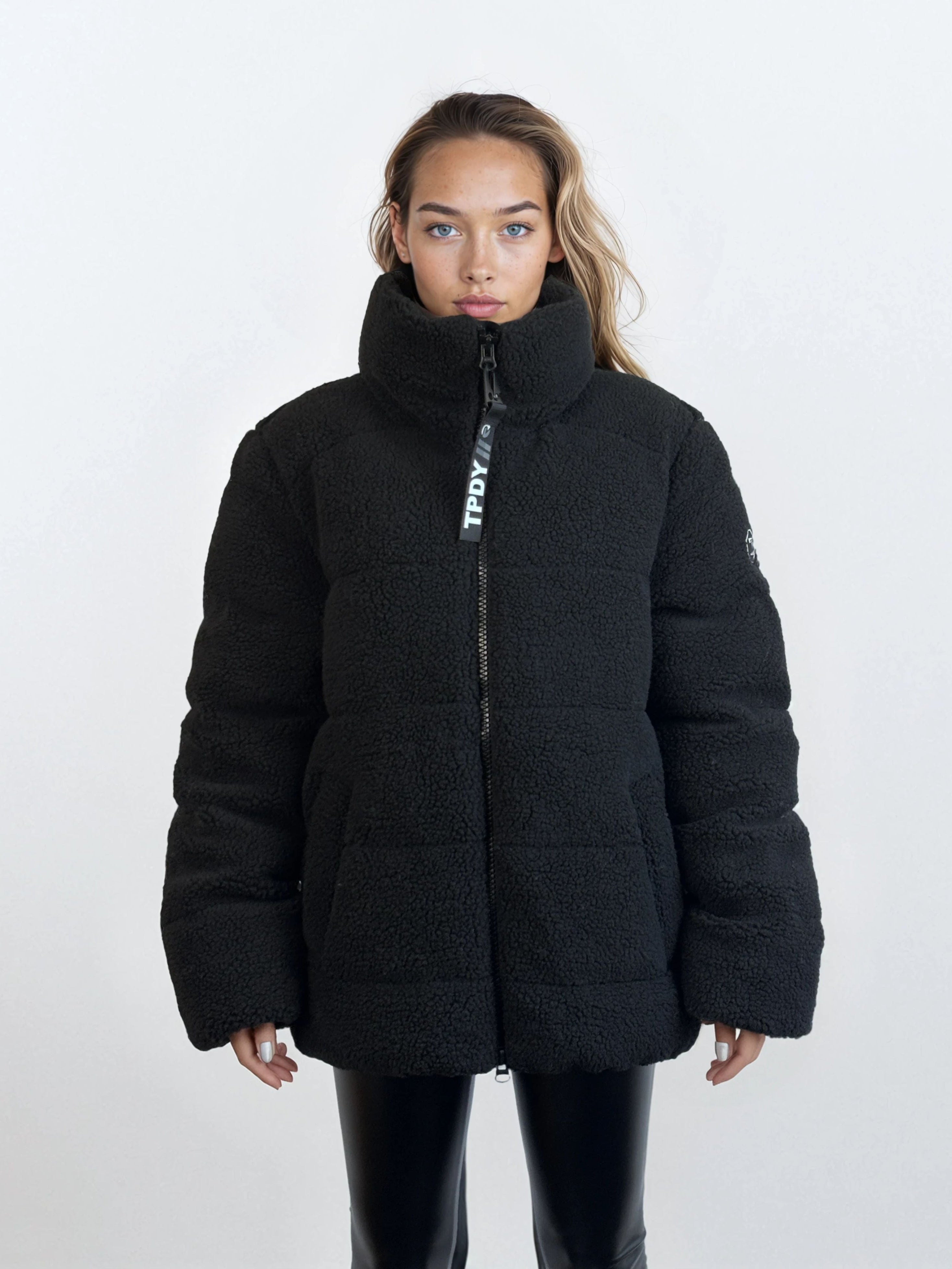 Eine Person steht mit dem Gesicht nach vorne und trägt eine schwarze, hochgeschlossene Pufferjacke mit Reißverschluss und eine schwarze Lederhose vor einem schlichten weißen Hintergrund.