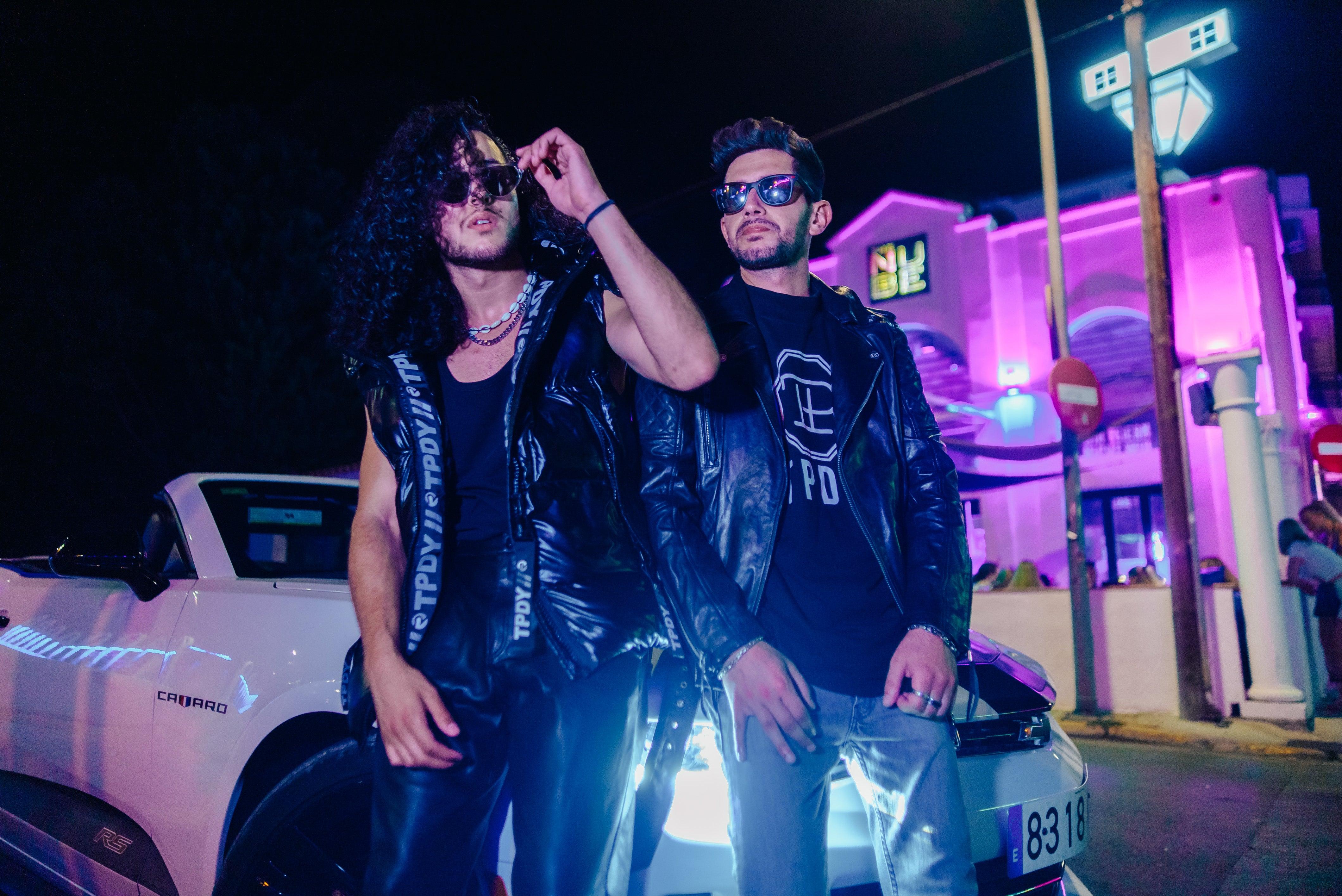 Zwei Männer mit modischer Lederjacke und Weste vor einem Cabrio. Im Hintergrund eine Diskothek bei Nacht