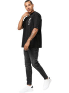 Ein Mann mit Tattoos auf dem linken Arm steht vor einem weißen Hintergrund und trägt das trueprodigy Jona Oversized T-Shirt mit Druck in schwarz, gepaart mit einer schwarzen Jeans und weißen Sneakers. Er blickt leicht nach links.