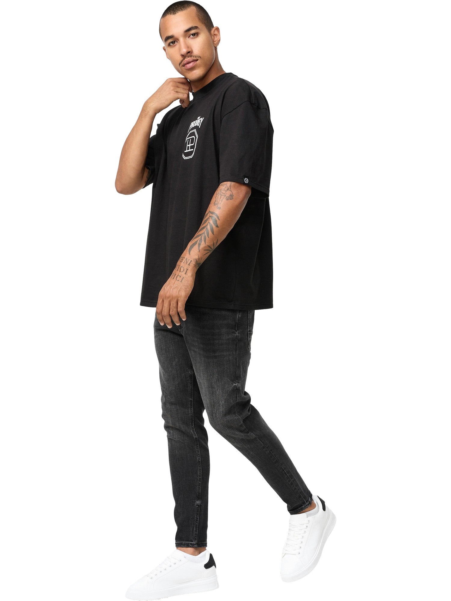 Ein Mann mit Tattoos auf dem linken Arm steht vor einem weißen Hintergrund und trägt das trueprodigy Jona Oversized T-Shirt mit Druck in schwarz, gepaart mit einer schwarzen Jeans und weißen Sneakers. Er blickt leicht nach links.