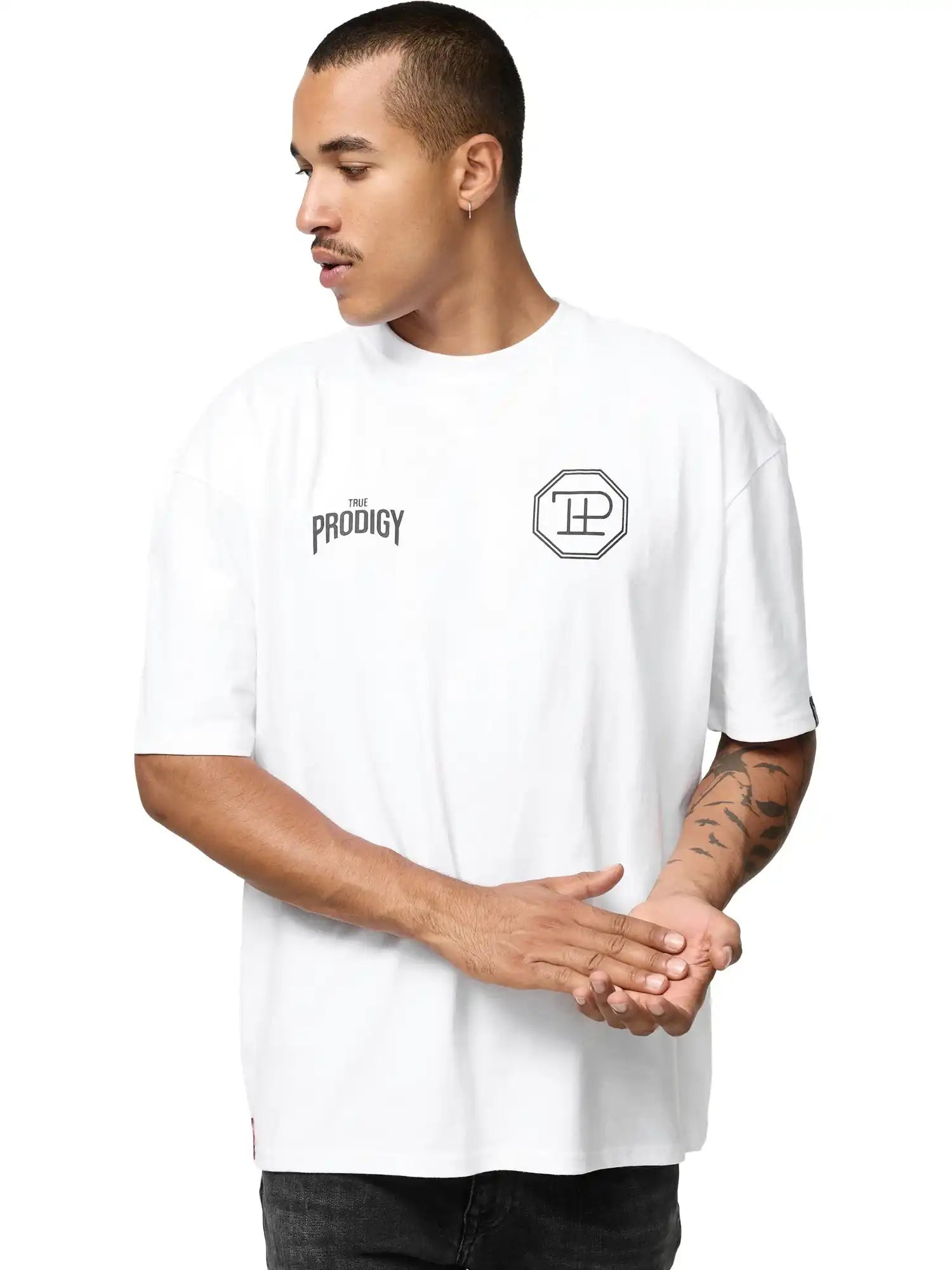 Eine Person trägt das Robin Oversized T-Shirt mit Druck von trueprodigy, ein weißes Unisex-Shirt mit THE PRODIGY-Schriftzug und einem achteckigen Logo auf der Vorderseite, steht mit zusammengelegten Händen und schaut zur Seite.