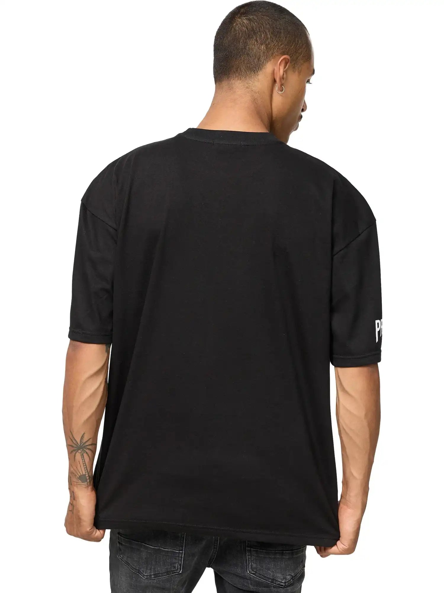 Eine Person mit kurzen Haaren und sichtbaren Tattoos am rechten Unterarm steht mit dem Rücken zur Kamera. Sie trägt das trueprodigy Dilan Oversized T-Shirt mit Druck und verwaschene schwarze Jeans.