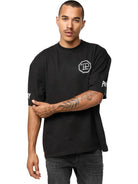 Eine Person mit kurzen Haaren und Schnurrbart trägt das trueprodigy Dilan Oversized T-Shirt mit Druck-schwarz, unisex, mit weißen grafischen Prints auf Brust und Ärmeln-gepaart mit dunklen Jeans, vor einem schlichten weißen Hintergrund.
