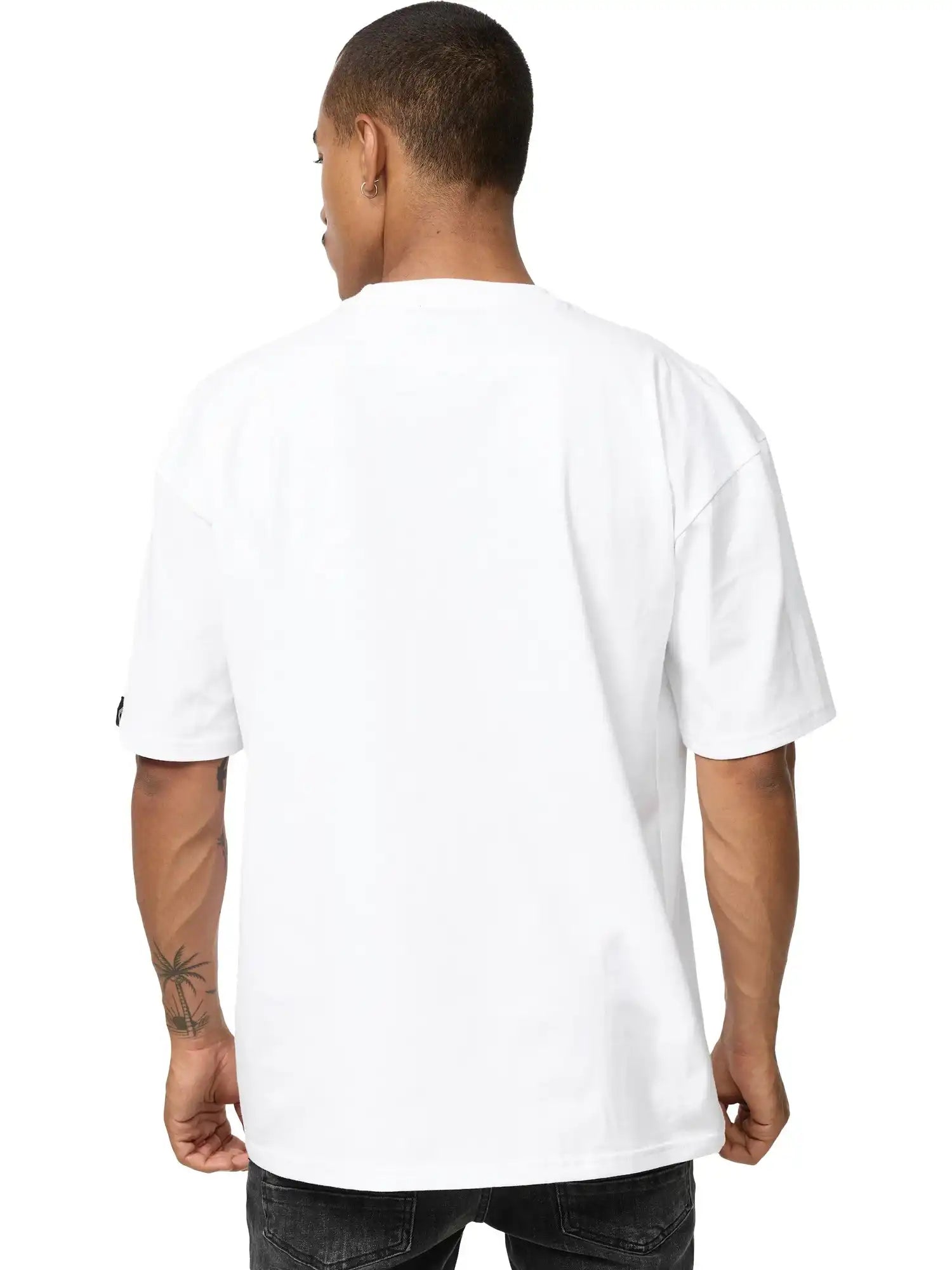 Eine Person mit kurzen Haaren und einer Tätowierung am linken Unterarm blickt von der Kamera weg und trägt ein schlichtes weißes trueprodigy Phoenix Oversized T-Shirt mit Stehkragen und dunkle Jeans.