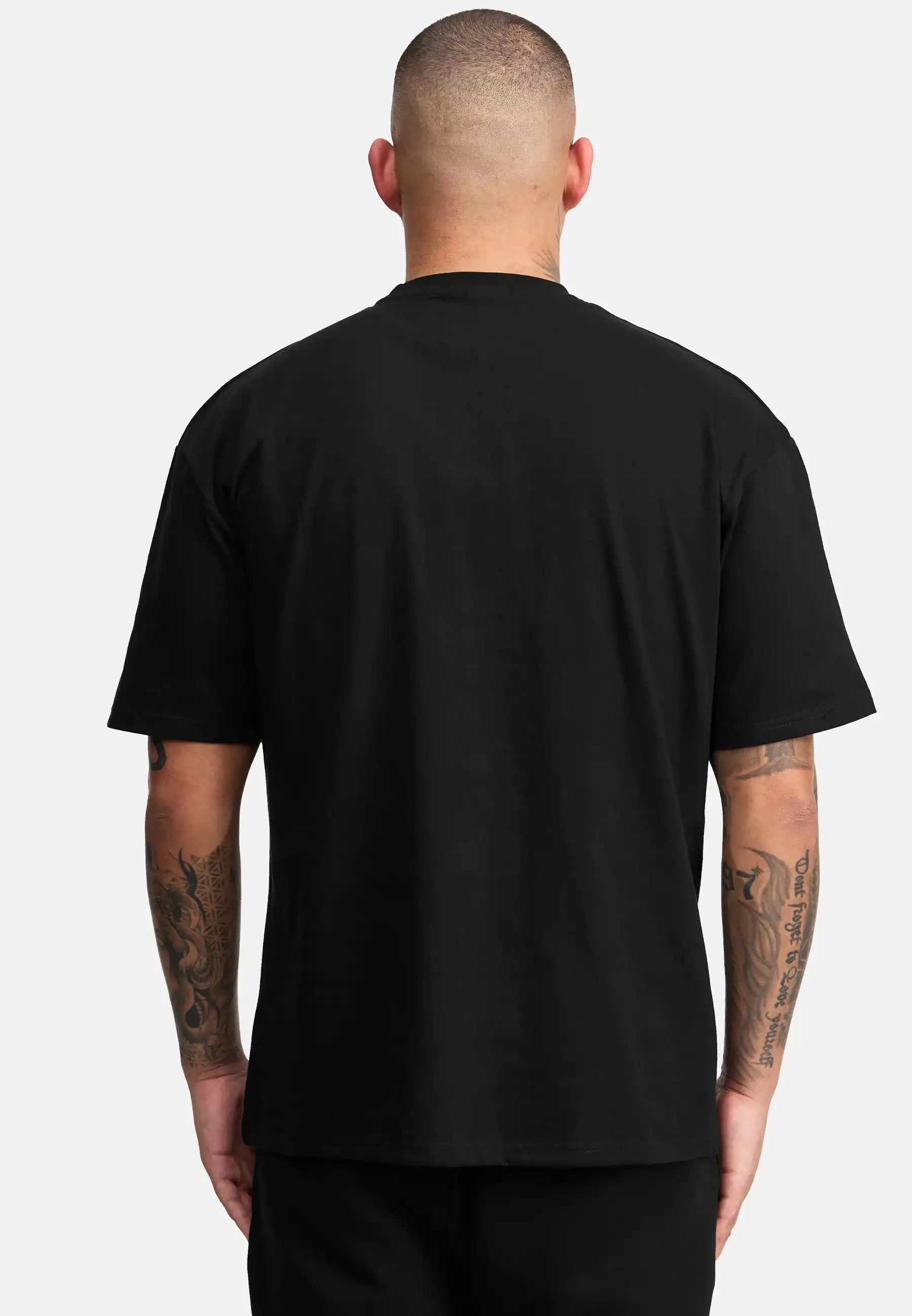 Eine Person mit kurzen Haaren ist von hinten zu sehen, die das trueprodigy Channing Oversized T-Shirt mit Druck trägt, gepaart mit einer schwarzen Hose. Beide tätowierten Arme sind sichtbar, und der schwere Jersey-Stoff hebt sich von einem hellgrauen Hintergrund ab.