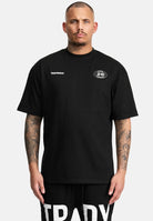 Ein Mann mit kurzen Haaren und Tattoos trägt das Channing Oversized T-Shirt mit Druck von trueprodigy - ein übergroßes Unisex-T-Shirt mit kleinen weißen Grafiken auf der Brust - und schwarze Shorts mit fetten weißen Schriftzügen vor einem schlichten hellen Hintergrund.