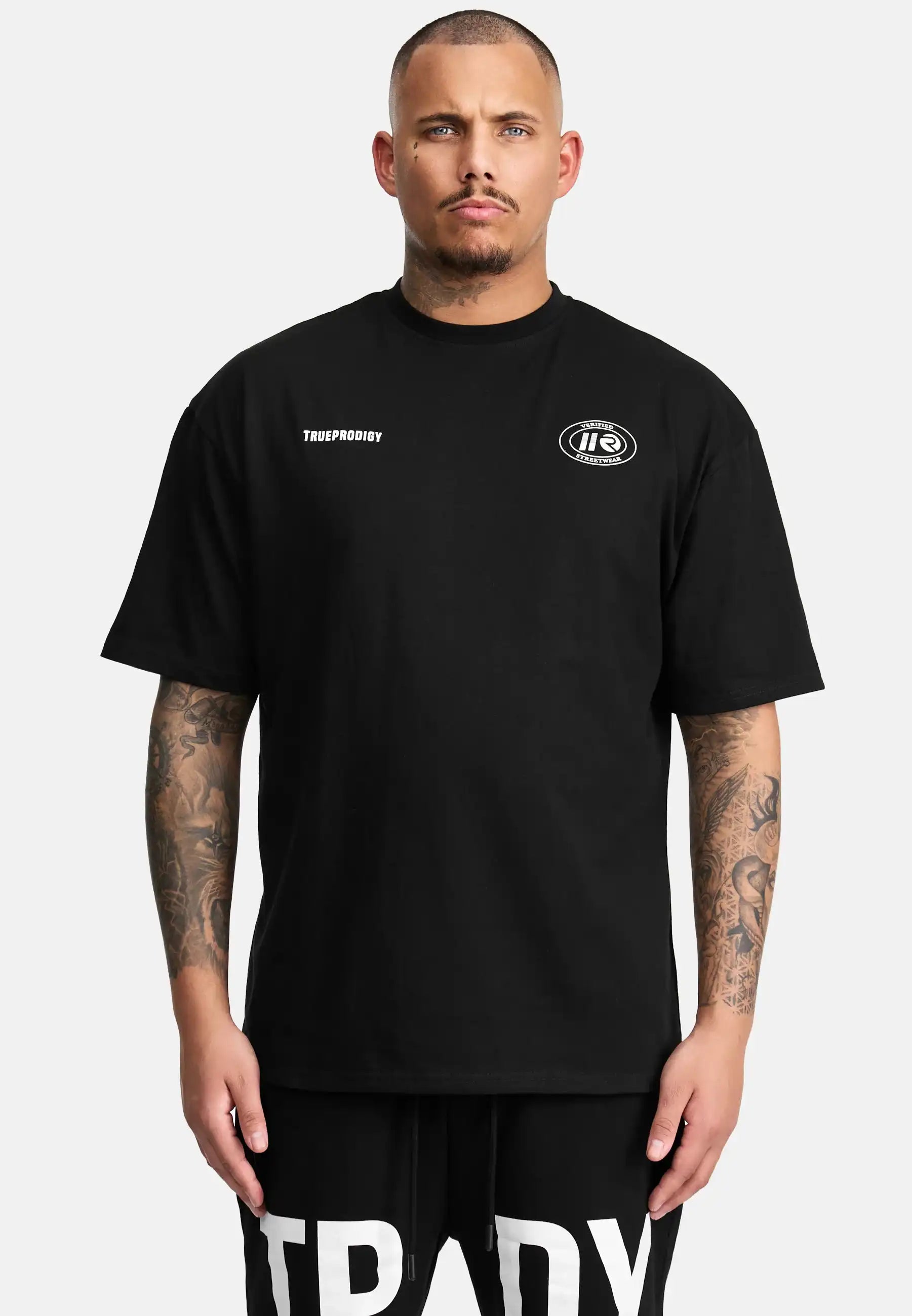 Ein Mann mit kurzen Haaren und Tattoos trägt das Channing Oversized T-Shirt mit Druck von trueprodigy - ein übergroßes Unisex-T-Shirt mit kleinen weißen Grafiken auf der Brust - und schwarze Shorts mit fetten weißen Schriftzügen vor einem schlichten hellen Hintergrund.