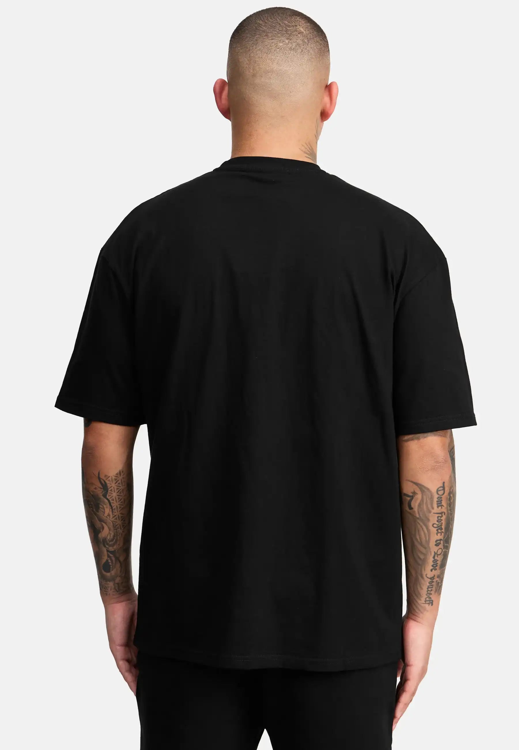 Eine Person mit kurzen Haaren und tätowierten Armen ist von hinten zu sehen. Sie trägt das trueprodigy Miguel Oversized T-Shirt mit Druck und eine schwarze Hose vor einem hellgrauen Hintergrund.