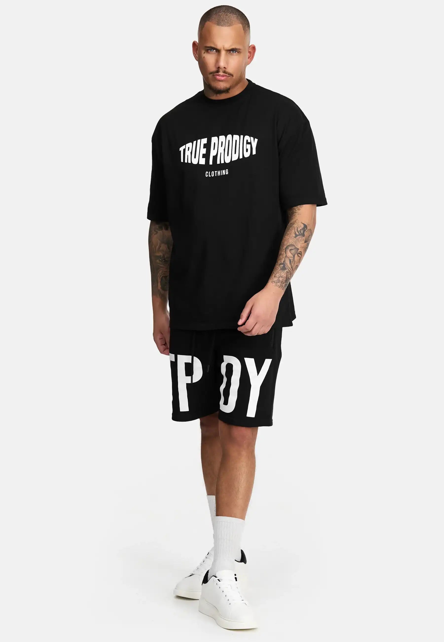 Ein Mann steht vor einem weißen Hintergrund und trägt das trueprodigy Miguel Oversized T-Shirt mit Druck, schwere schwarze Shorts mit großen weißen Buchstaben, weiße Socken und weiße Turnschuhe.