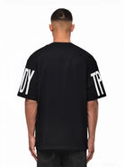 Ein Mann blickt von der Kamera weg und trägt das trueprodigy Viktor Oversize-Herren-T-Shirt mit schwerem Jersey-Stoff und großen weißen "TPD"- und "Y"-Ärmelaufdrucken vor einem schlichten weißen Hintergrund.