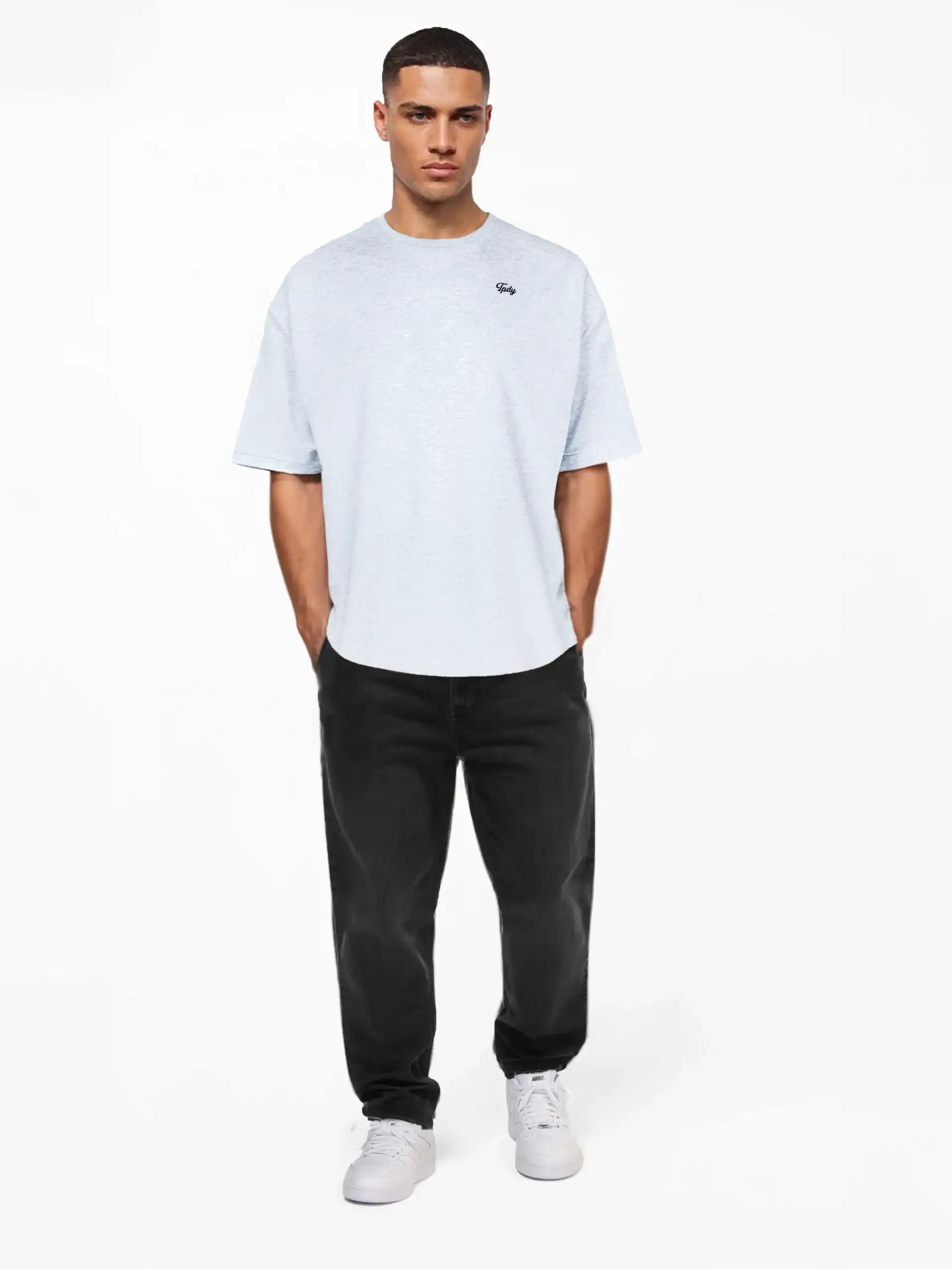 Ein Mann blickt nach vorne und trägt das trueprodigy Nika Oversize-Basicshirt mit TPDY-Stickerei & klarer Silhouette in Hellgrau, gepaart mit schwarzer Jeans und weißen Sneakers. Er steht vor einem schlichten weißen Hintergrund mit Händen in den Taschen und einem neutralen Gesichtsausdruck.
