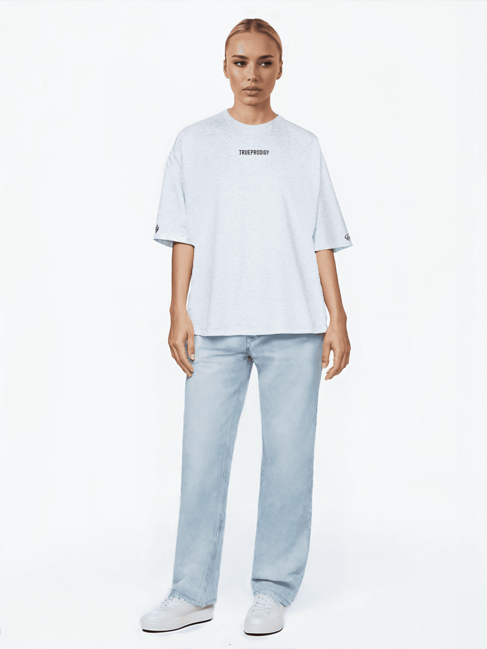Eine Person steht vor einem schlichten weißen Hintergrund und trägt ein hellgraues, übergroßes Unisex-T-Shirt „Kai F“ von trueprodigy mit der Aufschrift „TRUEPBGDRY“ auf der Vorderseite. Dazu trägt sie hellblaue Jeans und helle Sneakers und behält so einen neutralen Ausdruck.