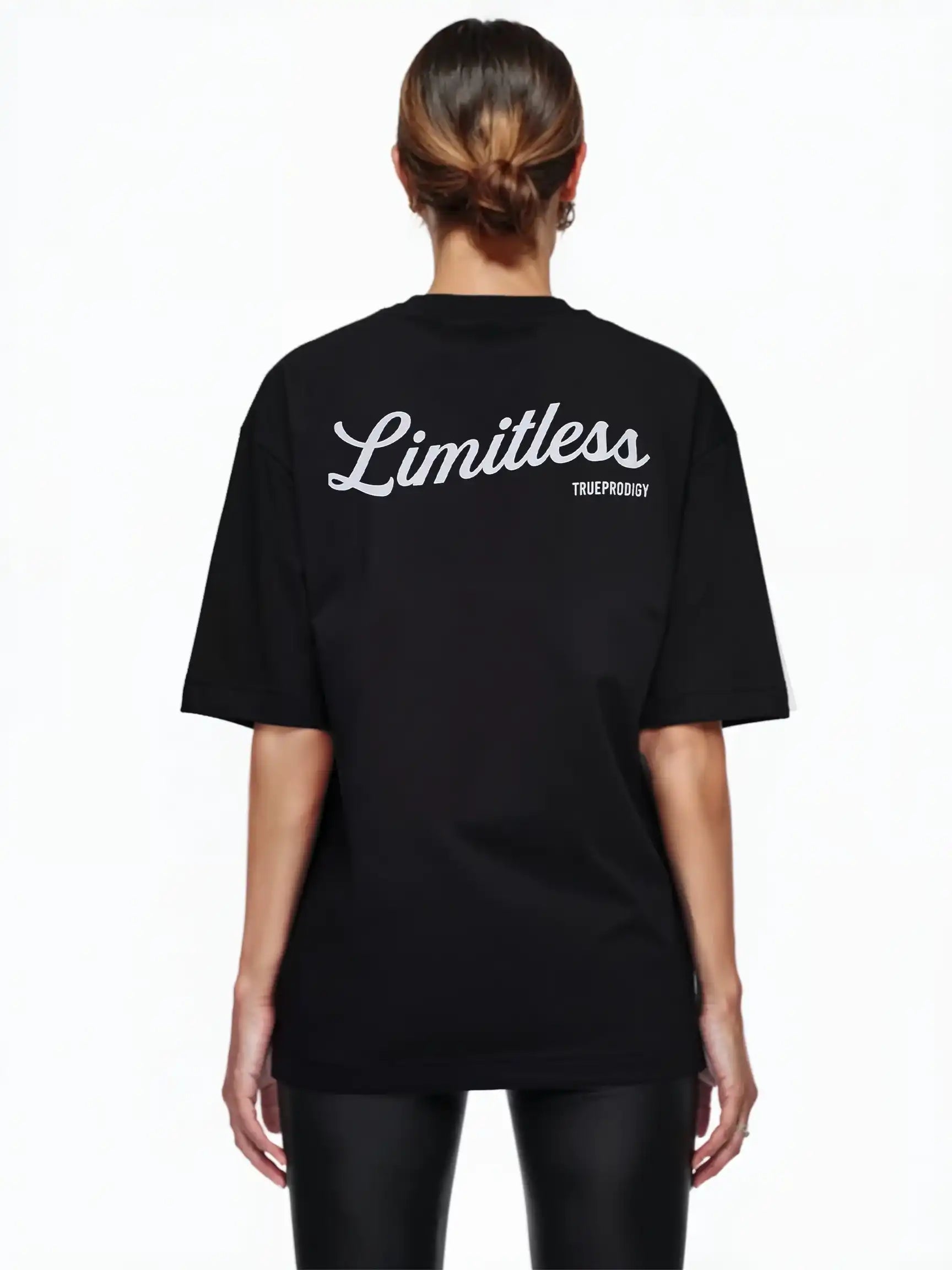 Eine Frau mit braunen Haaren in einem tiefen Dutt ist von hinten zu sehen. Sie trägt das Kai F Oversize-Damen-T-Shirt von trueprodigy mit einem "LIMITLESS"-Rückenaufdruck, gepaart mit einer schwarzen Hose vor einem schlichten weißen Hintergrund.