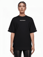 Vor weißem Hintergrund trägt eine Person ein übergroßes Unisex-T-Shirt mit dem auffälligen Aufdruck „trueprodigy“ auf der Vorderseite. Ihre hellen Haare sind hochgesteckt, und eine elegante schwarze Hose rundet den Look ab. (Produkt: Kai F von trueprodigy).