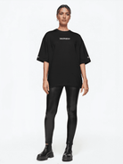 Eine Person steht vor einem weißen Hintergrund und trägt ein übergroßes Unisex-T-Shirt „Kai F“ mit dem „Trueprodigy“-Logo, glänzende schwarze Leggings und klobige Stiefel. Ihre Haare sind zurückgebunden, die Hände ruhen an den Seiten.