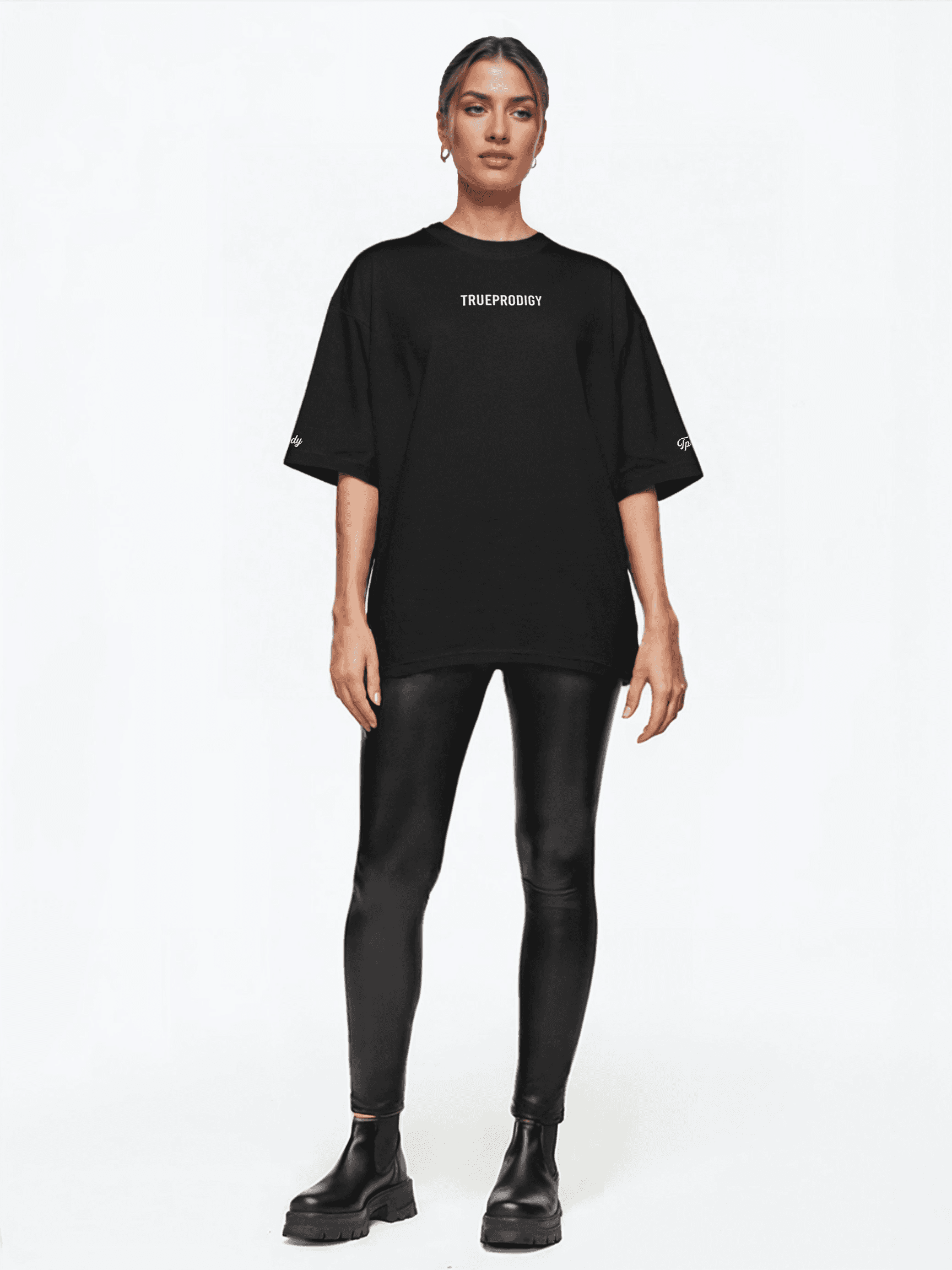 Eine Person steht vor einem weißen Hintergrund und trägt ein übergroßes Unisex-T-Shirt „Kai F“ mit dem „Trueprodigy“-Logo, glänzende schwarze Leggings und klobige Stiefel. Ihre Haare sind zurückgebunden, die Hände ruhen an den Seiten.