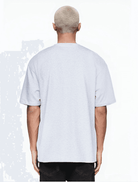 Eine Person mit kurzem, blondiertem Haar, die von der Kamera abgewandt ist, trägt ein übergroßes Trueprodigy Ravi Unisex-T-Shirt in Hellgrau mit dunkler Hose vor hellem Hintergrund.