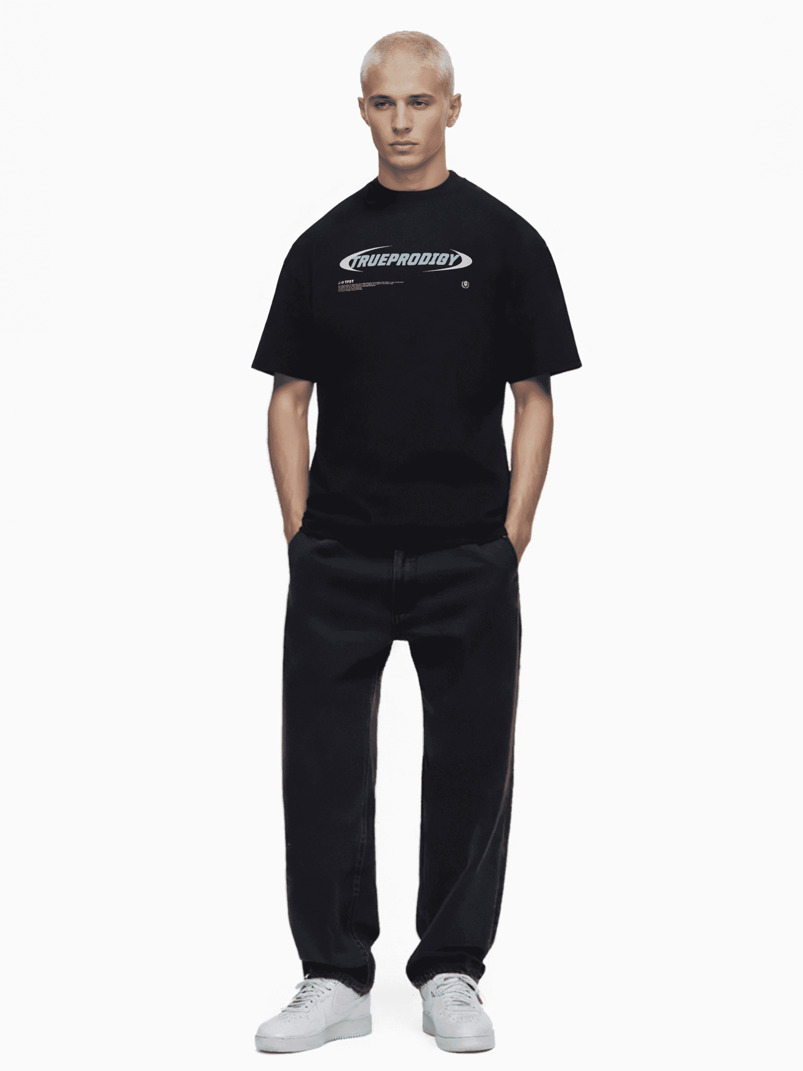 Eine Person mit kurzen blonden Haaren steht vor einem weißen Hintergrund und trägt ein Trueprodigy „Ravi“-Unisex-T-Shirt mit einem auffälligen blauen Logo. Dazu trägt sie eine schwarze Hose und weiße Turnschuhe, die Hände ruhen in den Taschen.