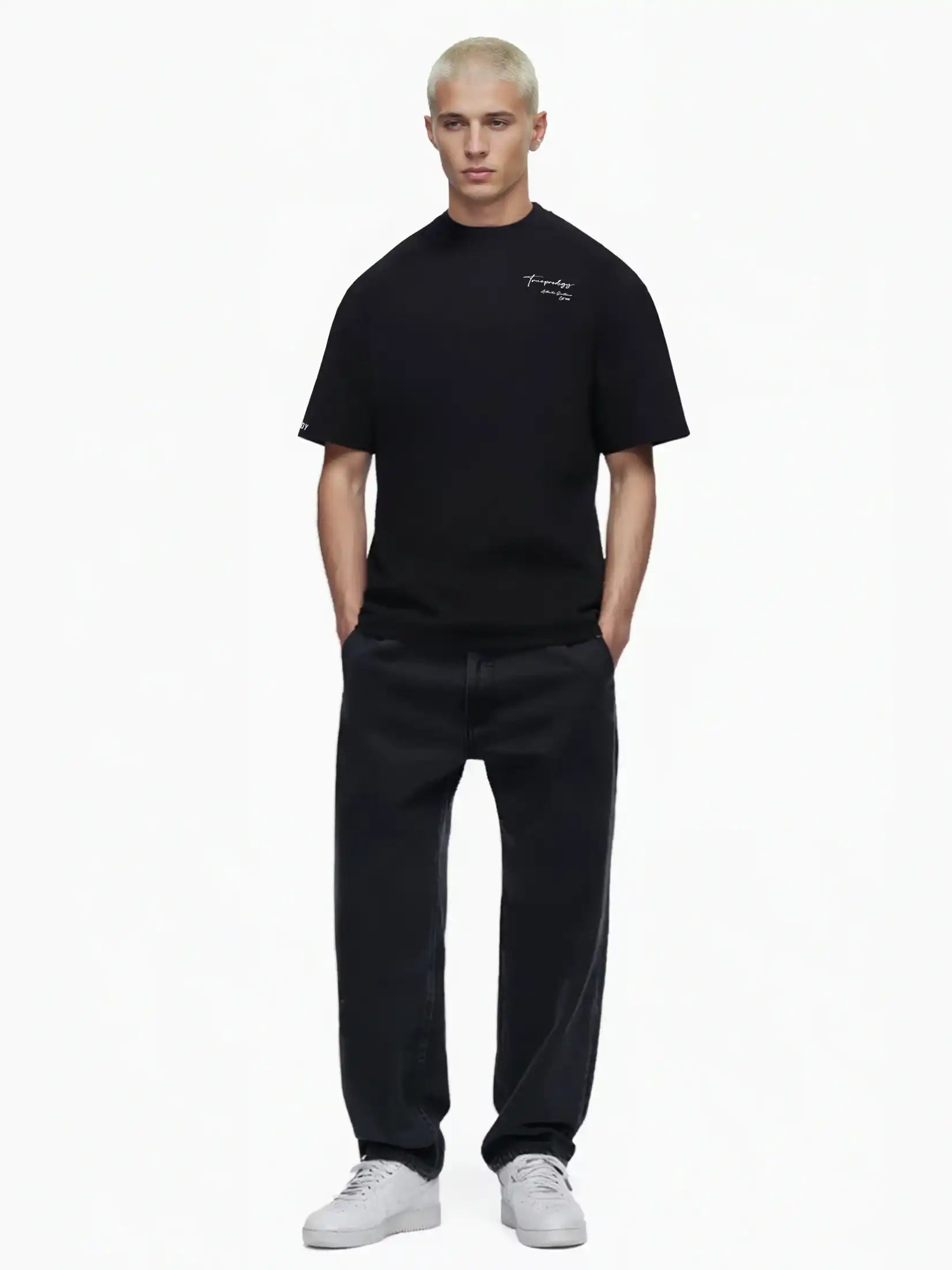 Eine Person mit kurzen blonden Haaren trägt das trueprodigy Marin Oversize-T-Shirt mit edlem Schriftzug auf der Brust, gepaart mit weiten schwarzen Jeans und weißen Turnschuhen, vor einem schlichten weißen Hintergrund.