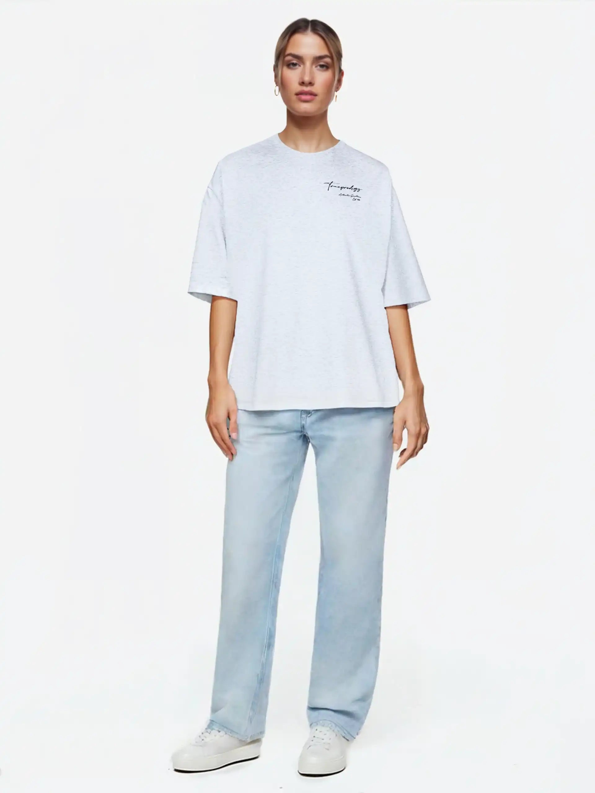 Eine Person schaut nach vorne und trägt ein hellgraues trueprodigy Marin F Oversize-T-Shirt mit feiner Logo-Schrift & TPDY-Print am Ärmel, gepaart mit hellblauen Jeans und weißen Turnschuhen, vor einem schlichten weißen Hintergrund.