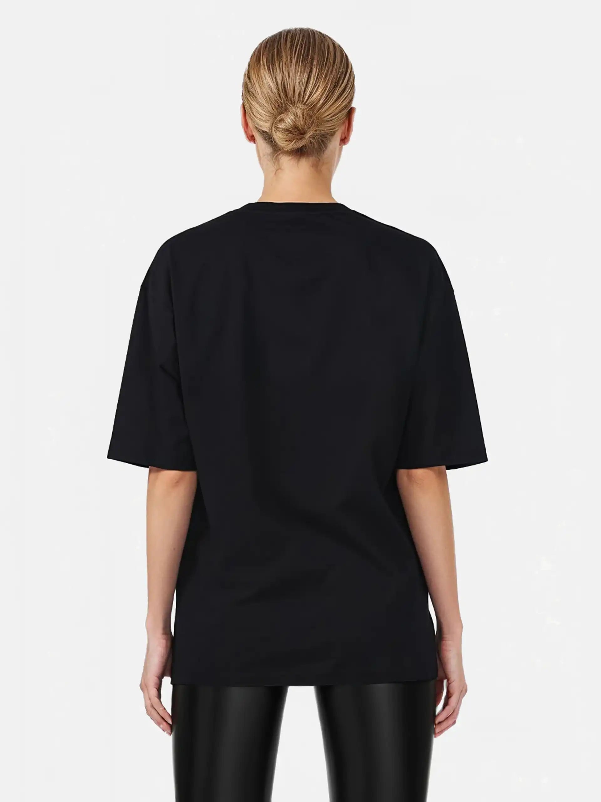 Eine Person mit blonden Haaren in einem tiefen Dutt ist von hinten zu sehen. Sie trägt das trueprodigy Marin F Oversize-T-Shirt mit feiner Logo-Schrift & TPDY-Print am Ärmel und eine schwarz glänzende Hose vor einem schlichten hellen Hintergrund.