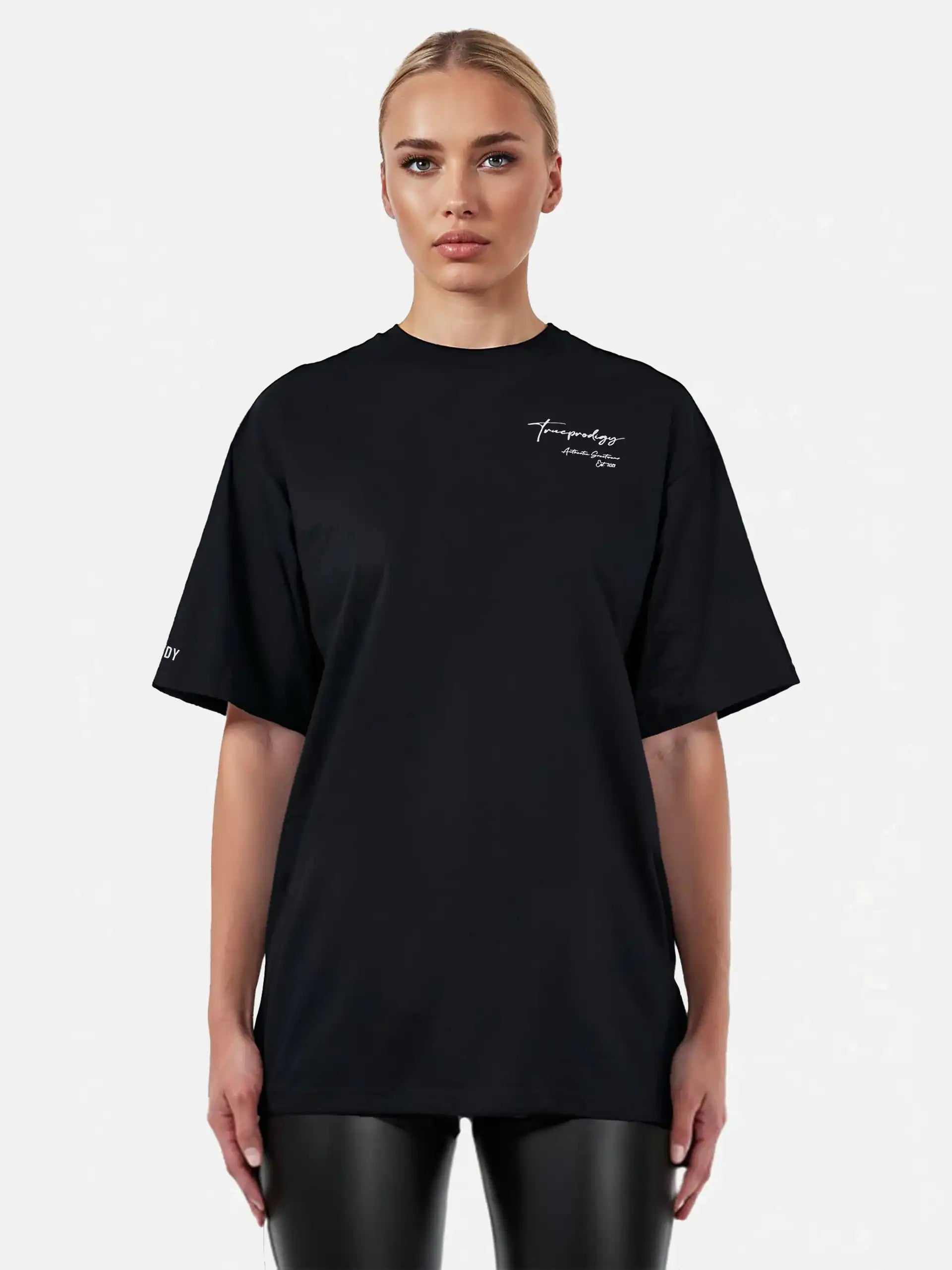 Eine Person blickt nach vorne in einem lockeren, unisex trueprodigy Marin F Oversize-T-Shirt mit feiner Logo-Schrift & TPDY-Print am Ärmel in Schwarz, gepaart mit einer schwarzen Lederhose, vor einem schlichten hellen Hintergrund.