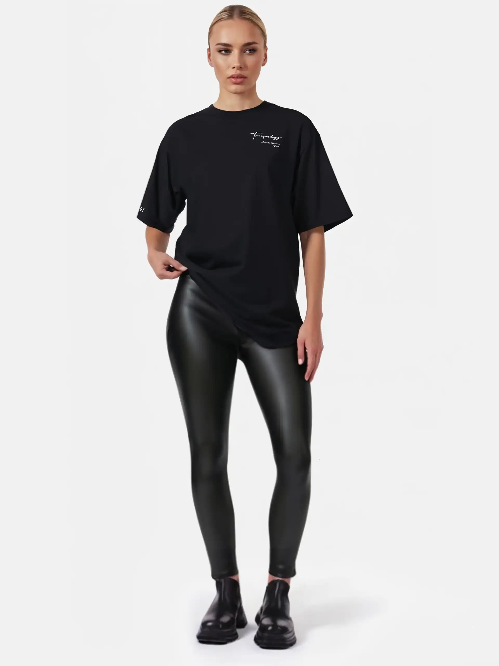 Eine Frau steht vor einem weißen Hintergrund und trägt das trueprodigy Marin F Oversize-T-Shirt mit feiner Logo-Schrift & TPDY-Print am Ärmel mit schwarz glänzenden Leggings und Schuhen. Ihr Haar ist zurückgesteckt und ihr Gesichtsausdruck ist neutral.