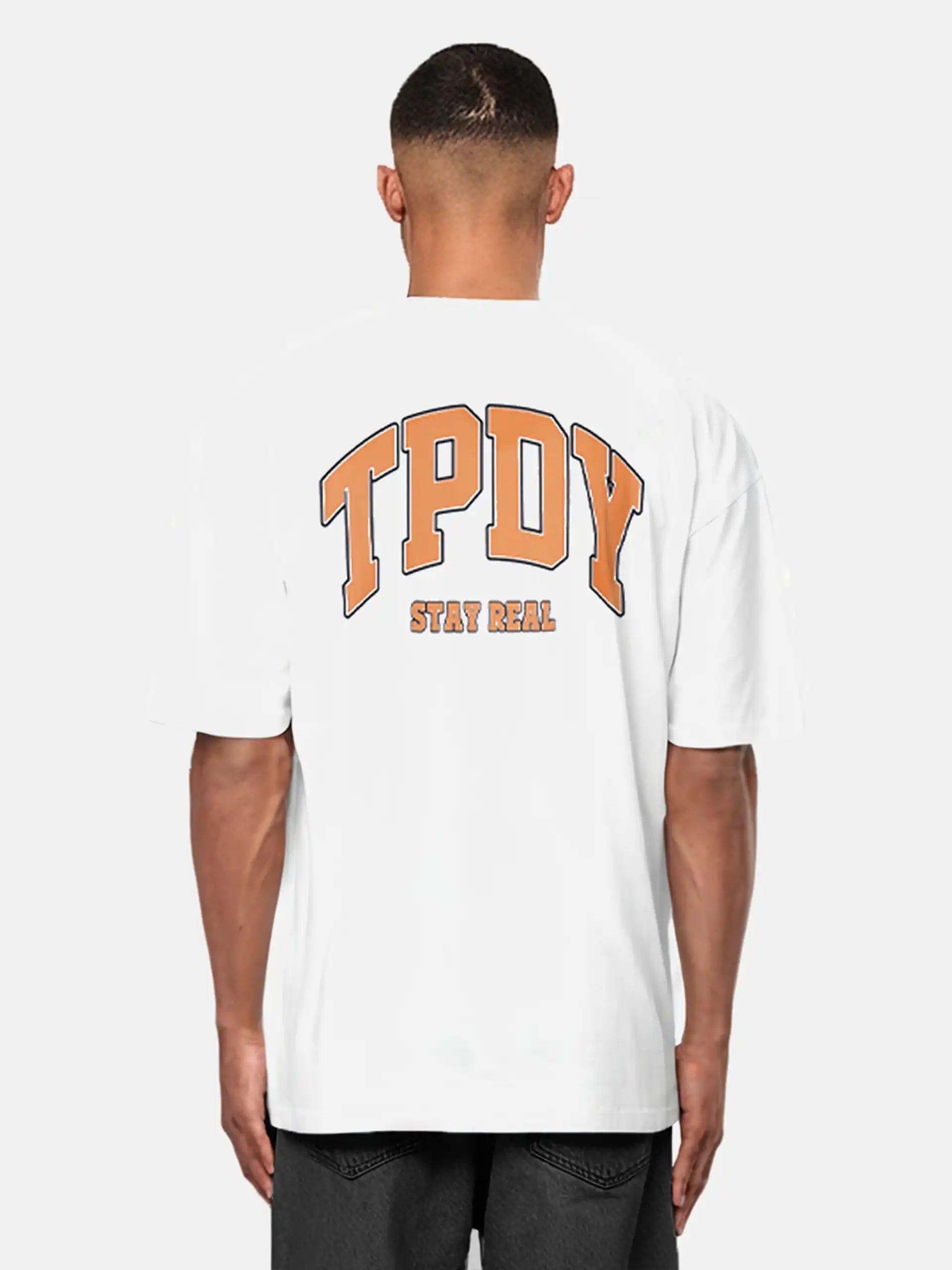 Jemand trägt das trueprodigy Oversize-Shirt mit TPDY-Logo & Rückenprint "Stay Real" - ein weißes Unisex-T-Shirt aus schwerem Jerseystoff mit auffälligem orangefarbenem TPDY-Schriftzug und "STAY REAL" am Rücken, vor hellem Hintergrund.