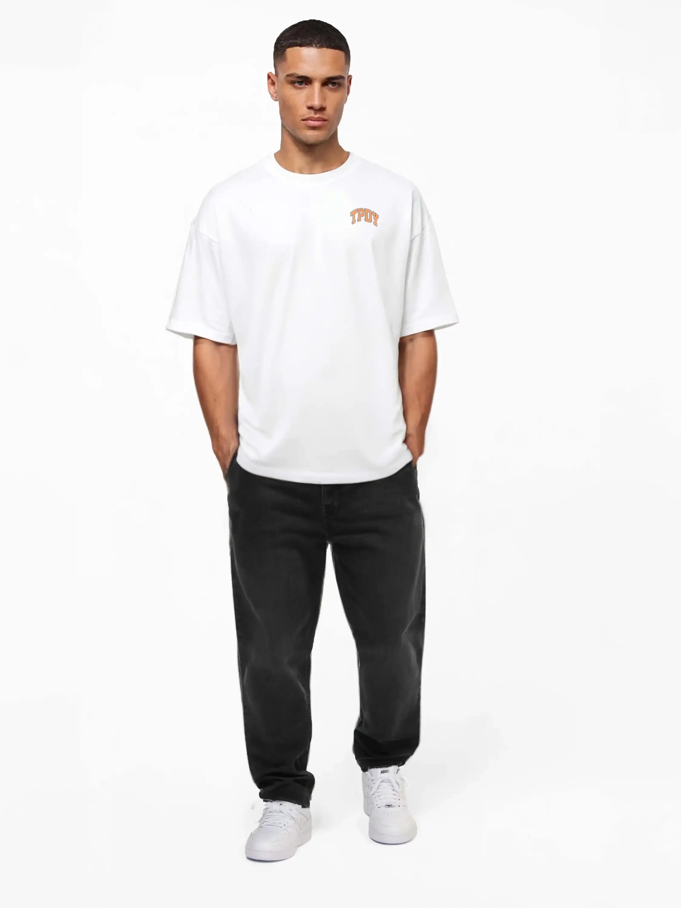 Ein Mann blickt nach vorne und trägt ein weißes trueprodigy Oversize-Shirt mit TPDY-Logo & Rückenprint "Stay Real", schwarze Jeans und weiße Turnschuhe. Er hat die Hände in den Taschen und steht vor einem schlichten weißen Hintergrund.