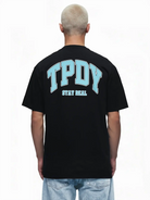 Eine Person mit kurzen, gebleichten Haaren ist von hinten zu sehen, die das trueprodigy Oversize-Shirt mit TPDY-Logo & Rückenprint "Stay Real" in Schwarz trägt, gestylt mit hellblauen Jeans.