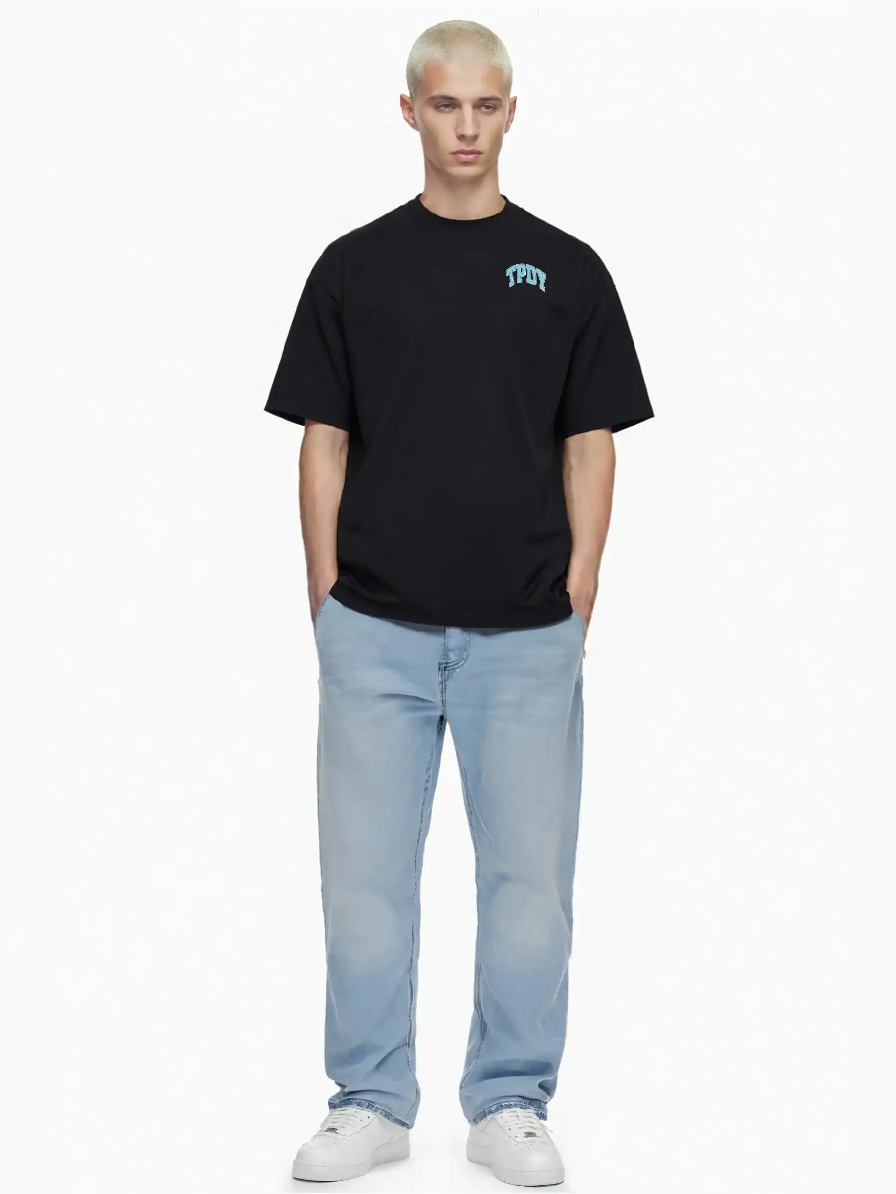 Eine Person mit kurzen, hellblonden Haaren steht vor einem weißen Hintergrund und trägt das trueprodigy Oversize-Shirt mit TPDY-Logo & Rückenprint "Stay Real", hellblaue Jeans und weiße Turnschuhe. Ihre Hände stecken in den Taschen.
