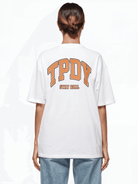 Eine Person trägt ein weißes Oversize-Unisex-T-Shirt „Arin F“ von trueprodigy mit dem „TPDY“- und „STAY REAL“-Logoprint in kräftigem Orange auf der Rückseite, perfekt kombiniert mit hellblauen Jeans. Ihre Haltung strahlt eine lässige und dennoch trendige Ausstrahlung aus, während sie von der Kamera abgewandt ist.