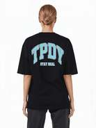 Eine blonde Person mit tiefem Dutt trägt ein übergroßes schwarzes Arin F-T-Shirt und eine dunkle Hose. Das Unisex-T-Shirt von trueprodigy trägt auf der Rückseite einen Logo-Aufdruck mit großen blauen Buchstaben „TPDY“ über dem Satz „STAY REAL“.