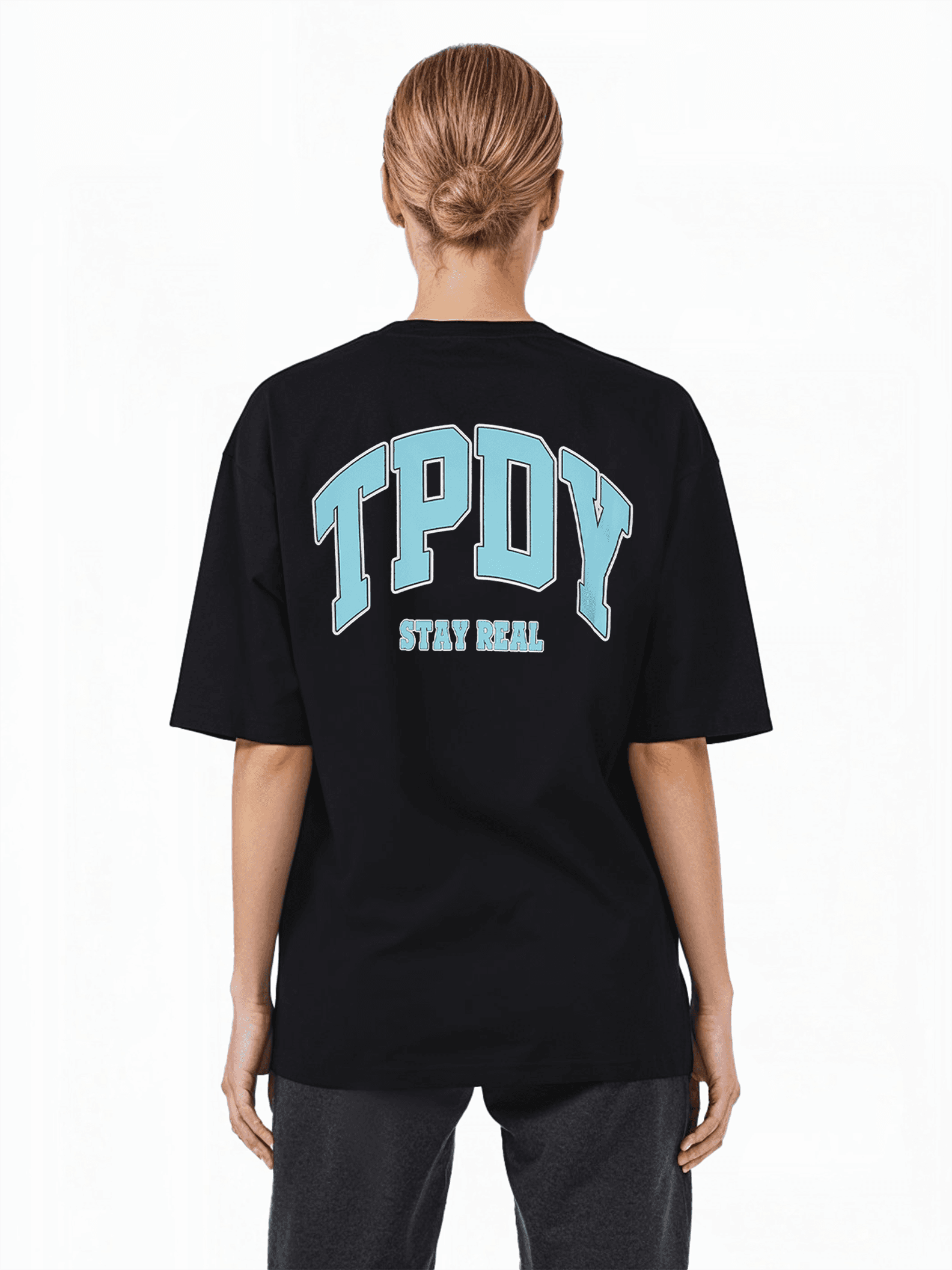 Eine blonde Person mit tiefem Dutt trägt ein übergroßes schwarzes Arin F-T-Shirt und eine dunkle Hose. Das Unisex-T-Shirt von trueprodigy trägt auf der Rückseite einen Logo-Aufdruck mit großen blauen Buchstaben „TPDY“ über dem Satz „STAY REAL“.