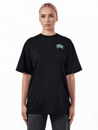 Eine Person mit kurzen blonden Haaren trägt das übergroße schwarze Arin F-T-Shirt von trueprodigy mit einem kleinen blauen „TPOY“-Logo auf der Vorderseite und eine dunkle Hose. Sie steht vor einem schlichten weißen Hintergrund und blickt direkt in die Kamera.