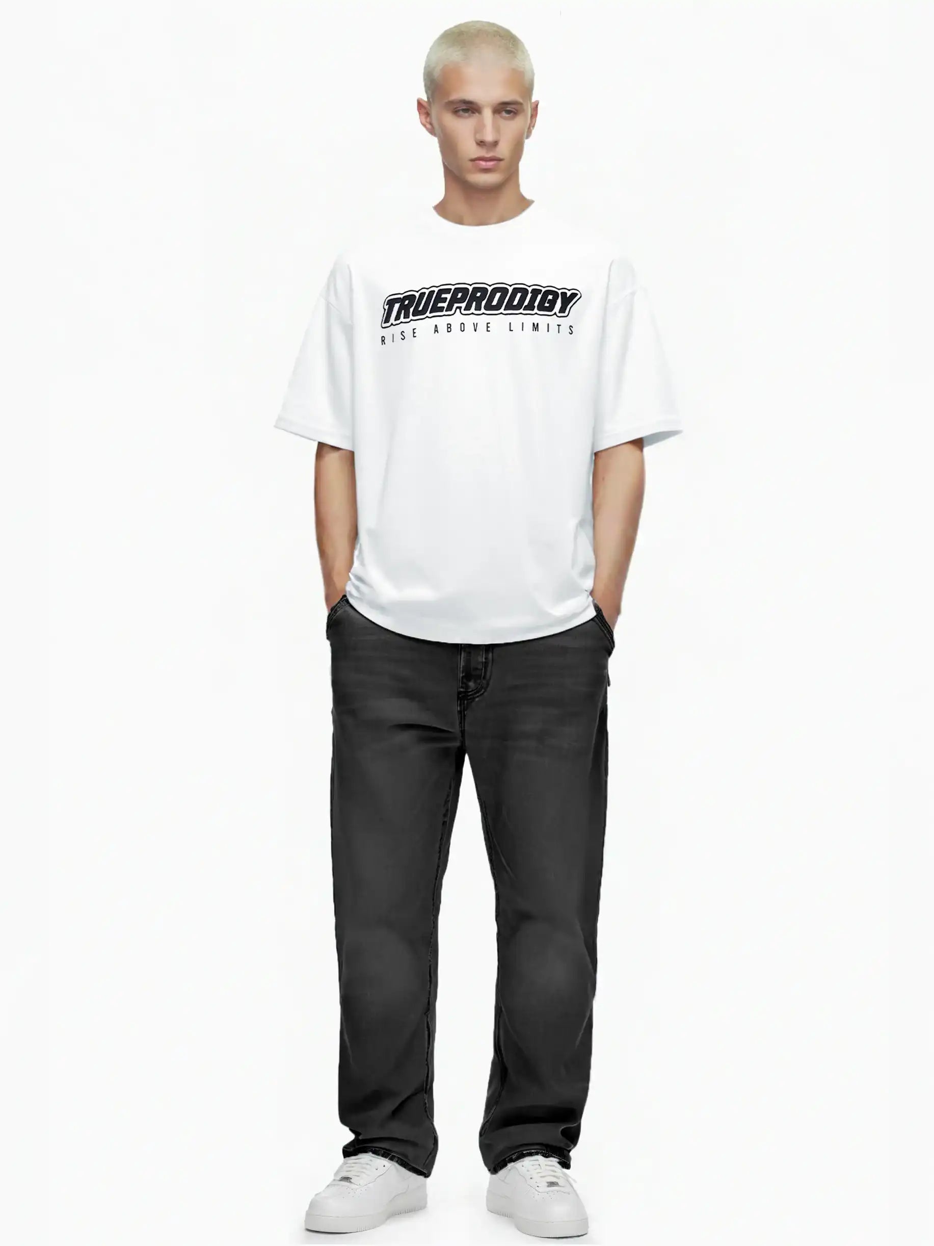 Eine Person mit kurzen blonden Haaren steht mit dem Blick nach vorne, trägt das trueprodigy Toni Oversize-Shirt mit 3D-Schaumprint & "Rise Above Limits", schwarze Jeans und weiße Turnschuhe. Ihre Hände stecken in den Taschen vor einem schlichten weißen Hintergrund.