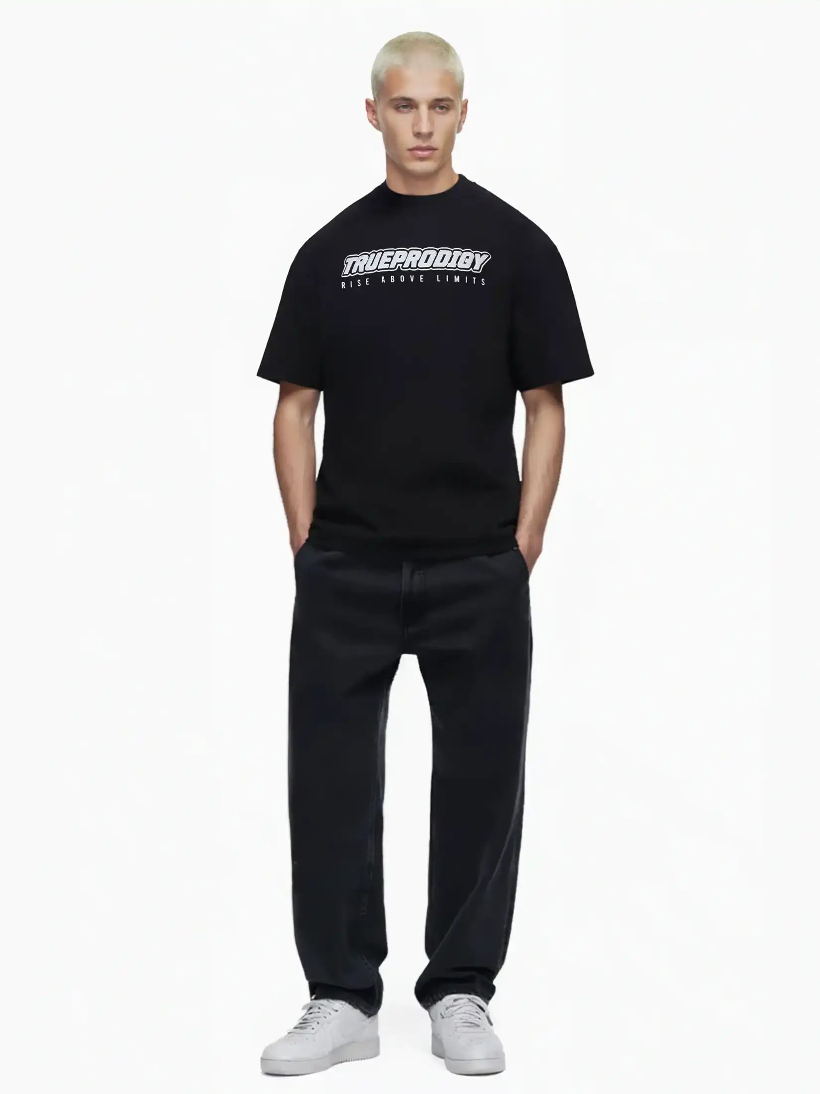Eine Person mit kurzen, gebleichten blonden Haaren trägt das trueprodigy Toni Oversize-Shirt mit 3D-Schaumprint & "Rise Above Limits", lockere schwarze Jeans und weiße Turnschuhe, stehend vor einem schlichten weißen Hintergrund.