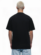 Eine Person mit kurzen, hellen Haaren steht mit dem Rücken zur Kamera, trägt ein schlichtes schwarzes trueprodigy Oversize-Herren-T-Shirt mit Logo-Smile-Grafik auf der Brust und hellblaue Jeans vor einem weißen Hintergrund.