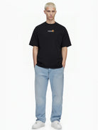 Eine Person mit kurzen blonden Haaren trägt das trueprodigy Oversize-Herren-T-Shirt mit Logo-Smile-Grafik auf der Brust, hellblaue Jeans und weiße Turnschuhe und steht mit den Händen in den Taschen vor einem weißen Hintergrund.