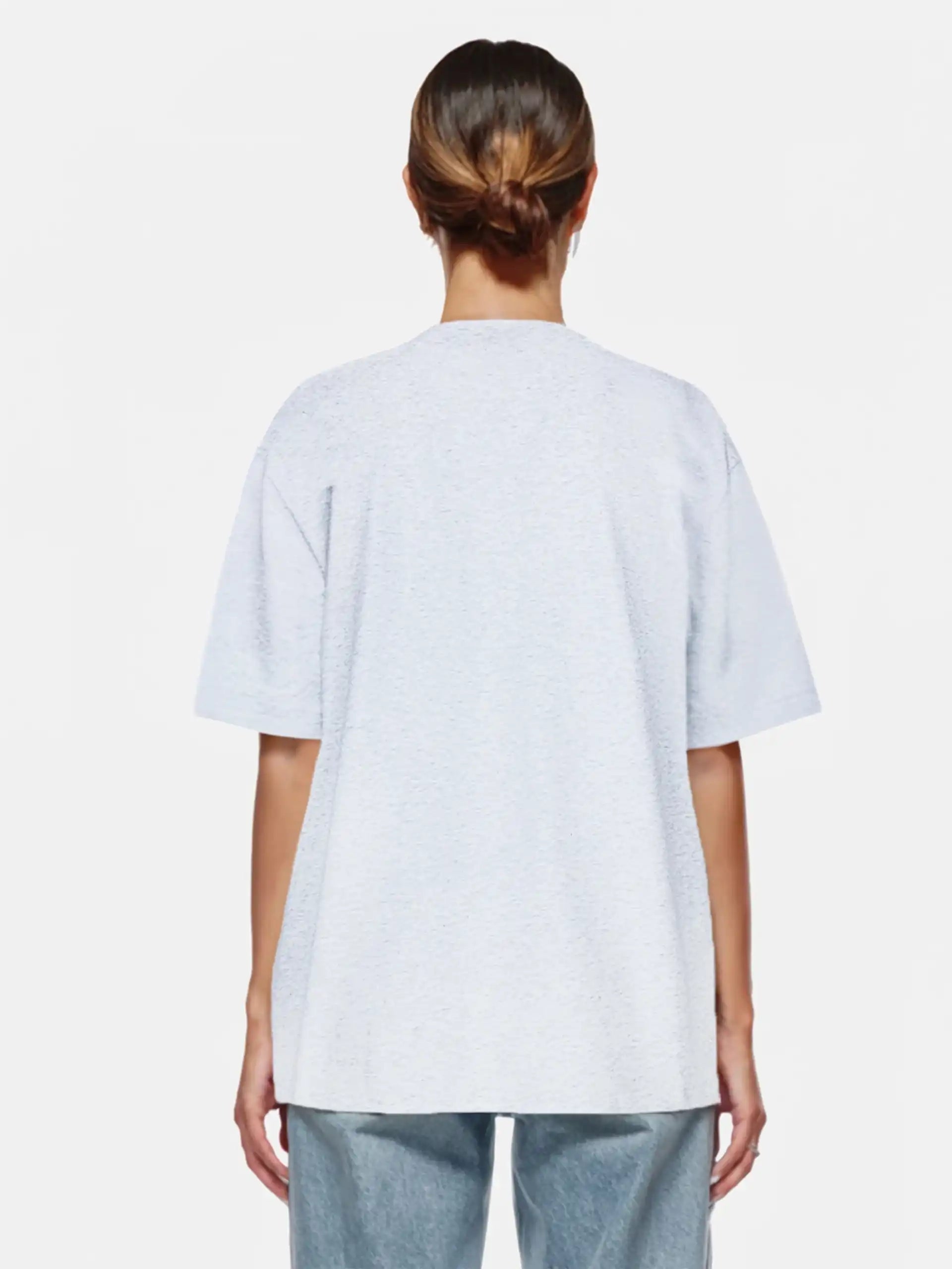 Eine Person mit braunen Haaren in einem tiefen Dutt steht mit dem trueprodigy Juniper F Oversize-Damen-T-Shirt mit Logo-Smile-Grafik und hellblauen Jeans vor einem schlichten weißen Hintergrund.
